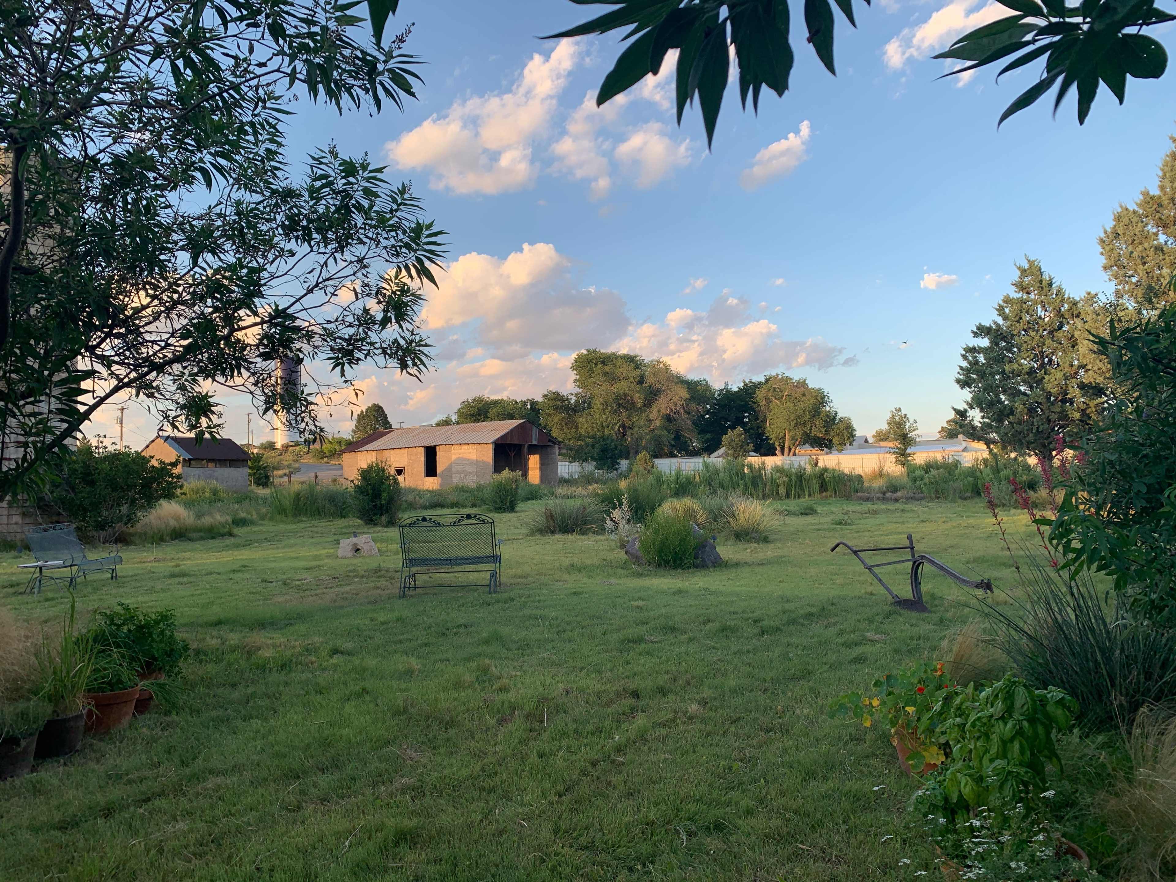 The image shows a peaceful outdoor area with grass, a few benches, and several trees under a partly cloudy sky.