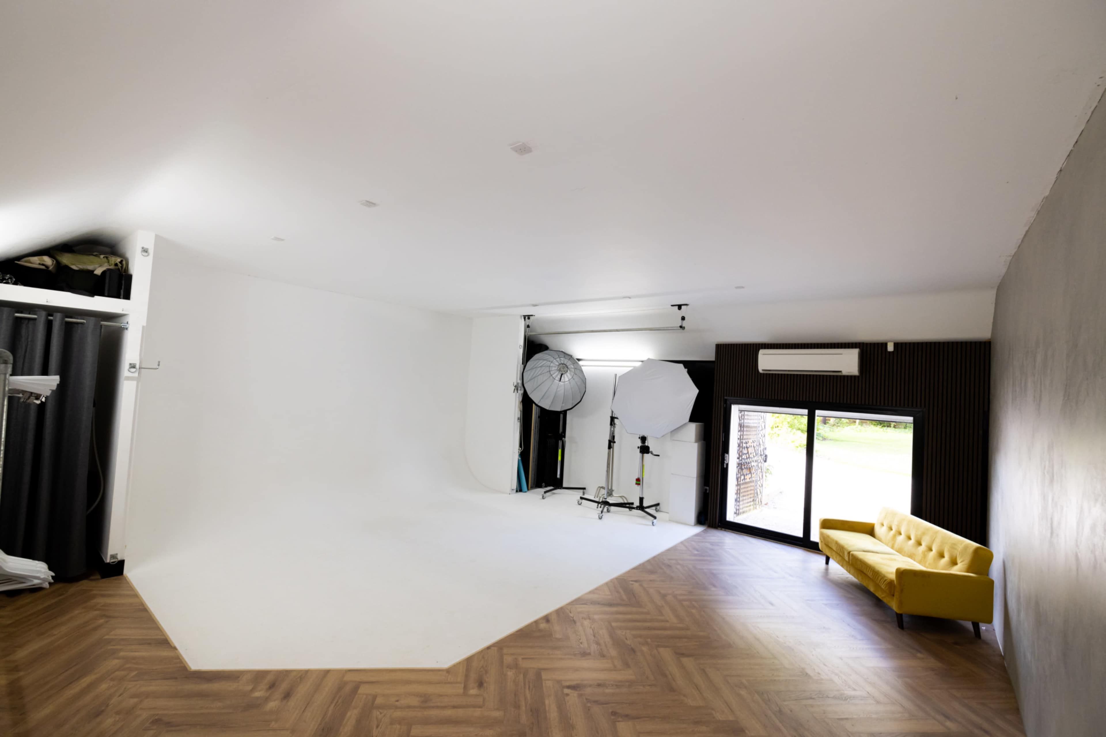 A spacious photography studio with a large white backdrop, two lighting setups, and a yellow couch against a dark wall.