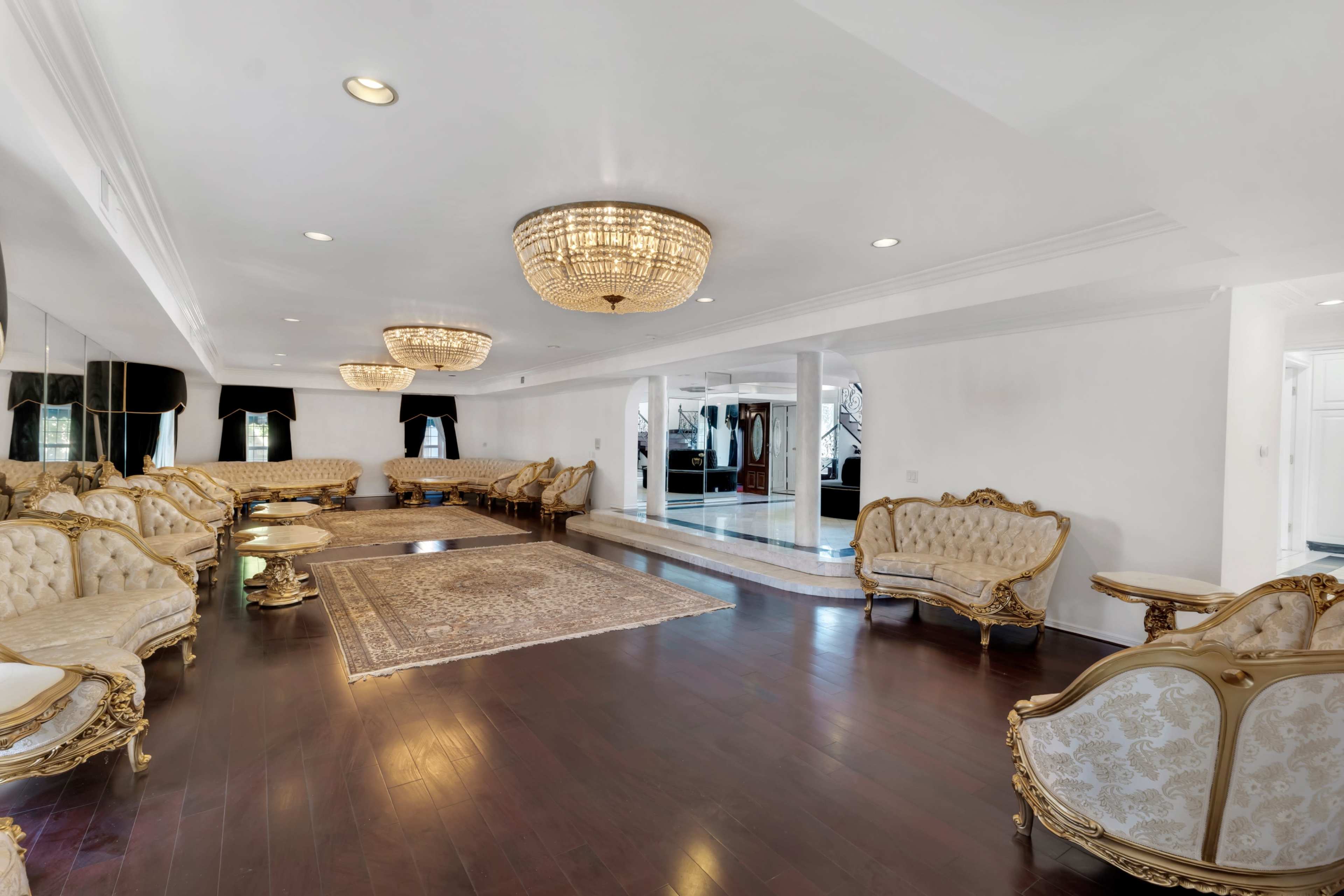 The image shows a spacious, elegantly furnished living room with ornate gold-framed furniture, two large chandeliers, and a hardwood floor.