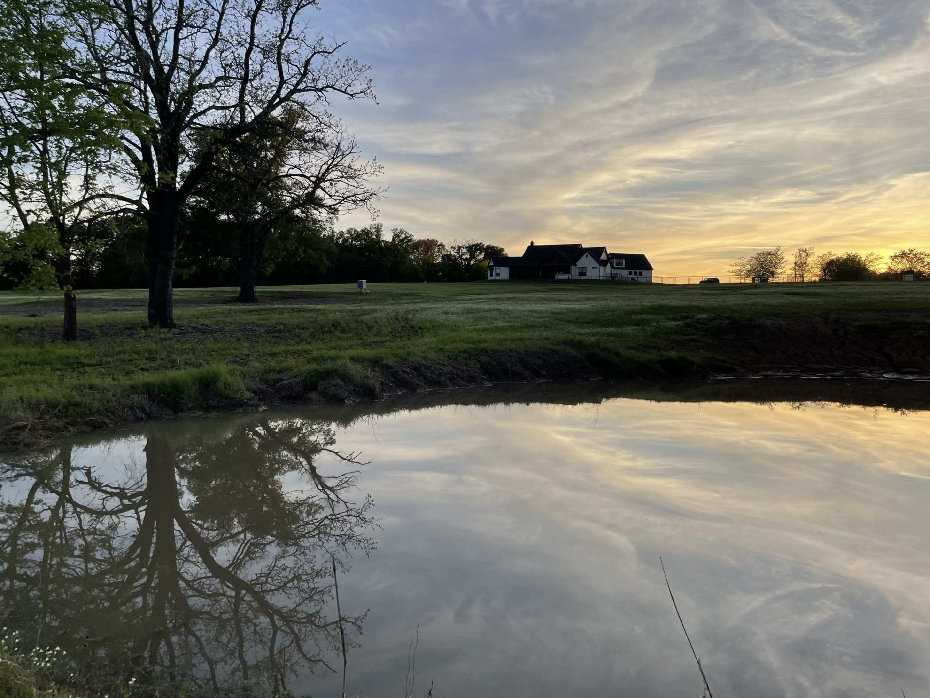 A house is situated near a pond that reflects the surrounding trees and the evening sky.