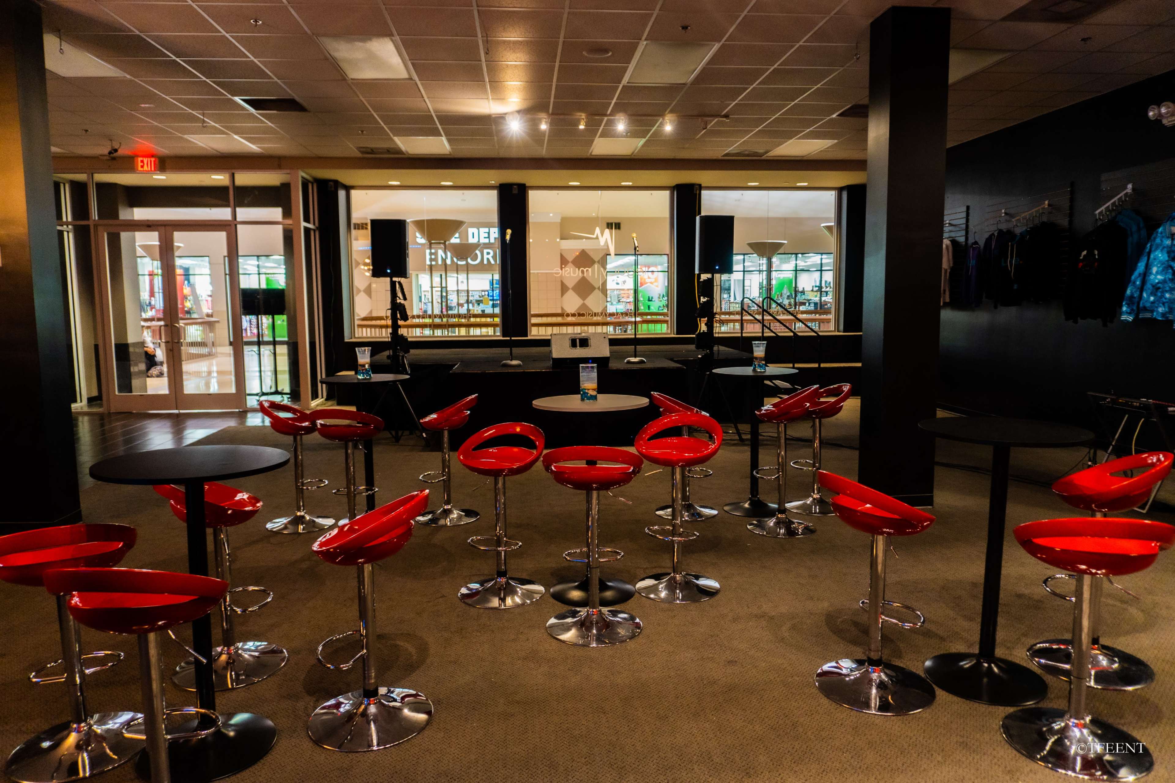 A spacious indoor venue with red bar stools arranged around small black tables, facing a glass-fronted area with displays.