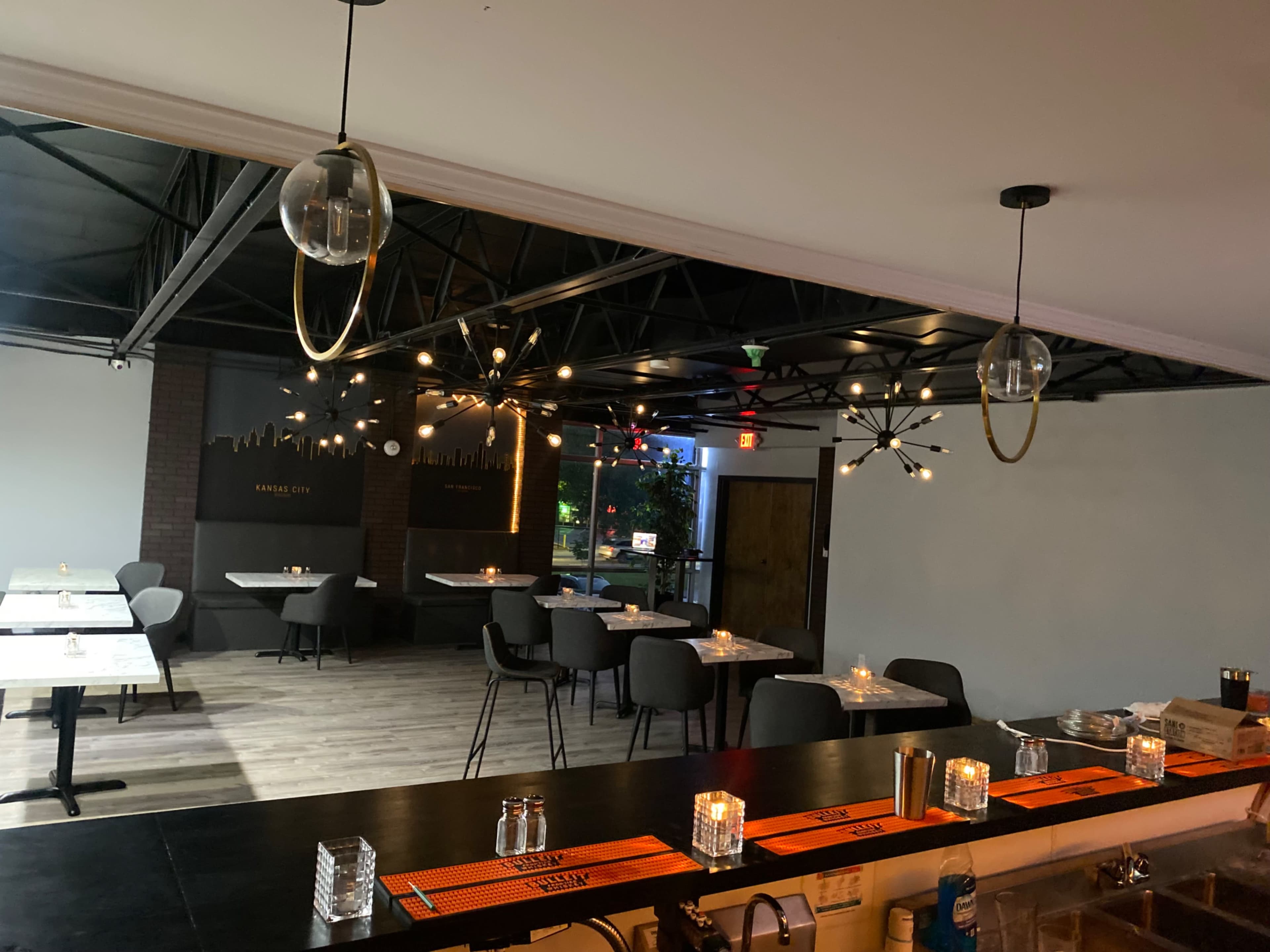 The image shows a modern restaurant interior with empty tables, a well-lit bar area, and decorative lighting fixtures hanging from the ceiling.