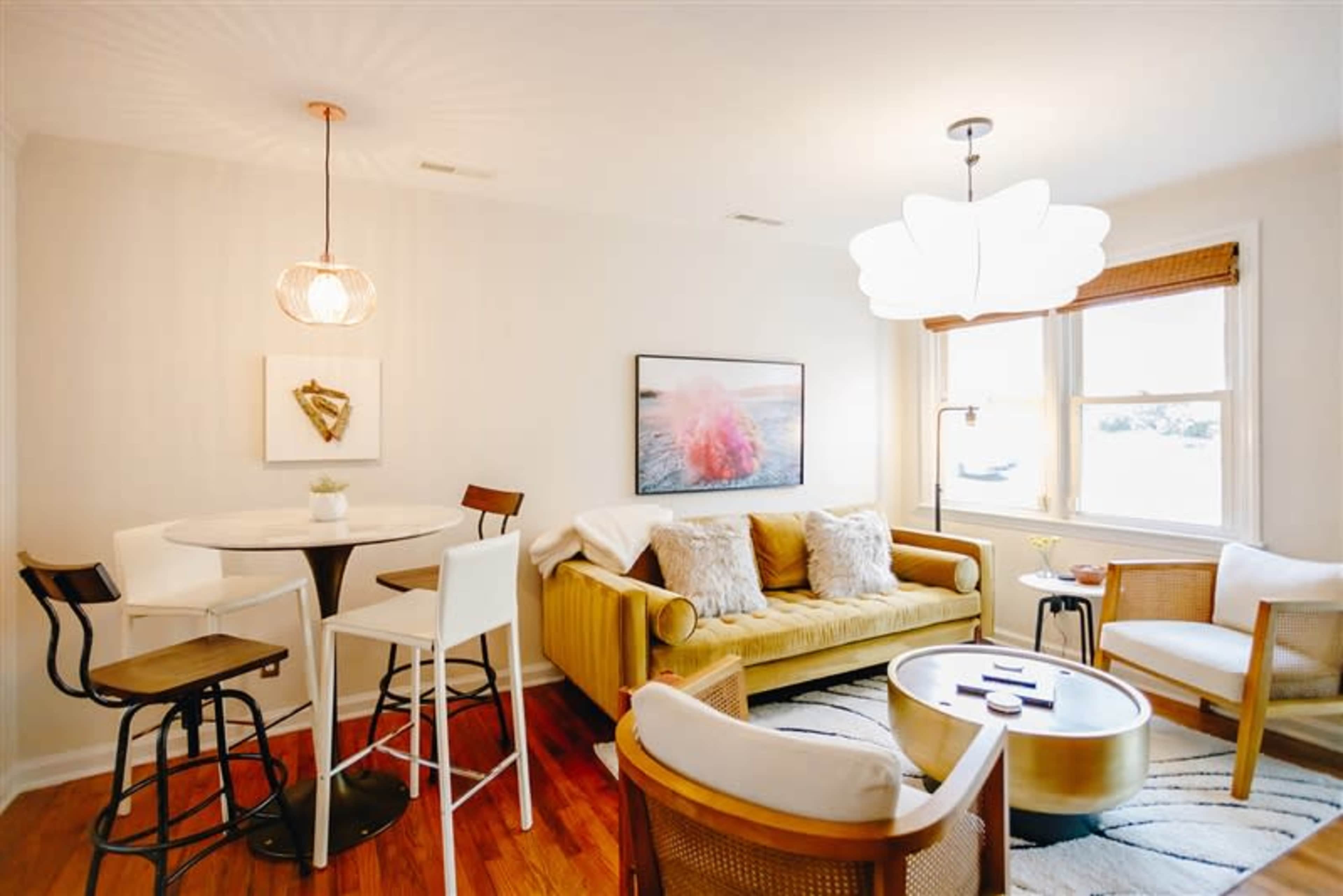 A cozy living area features a yellow sofa, three seating chairs, a round table with bar stools, and large windows providing ample light.