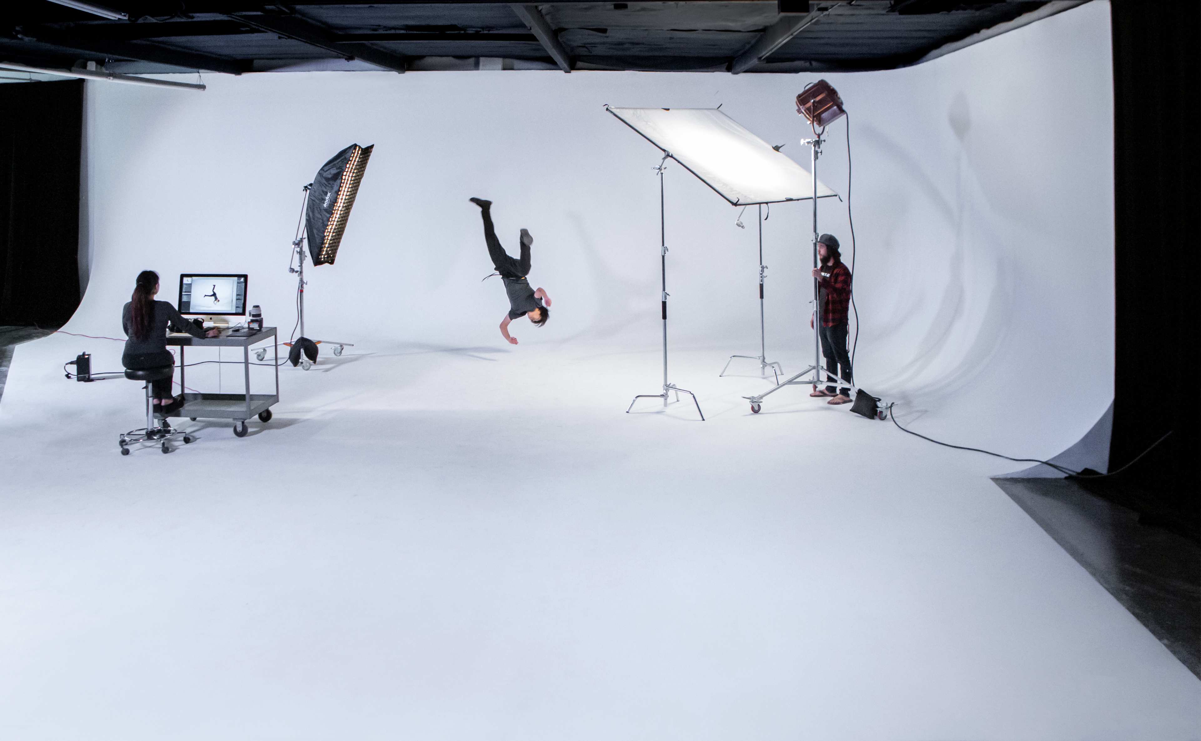 A dancer performs a handstand in a photography studio with lighting equipment and a computer workstation.