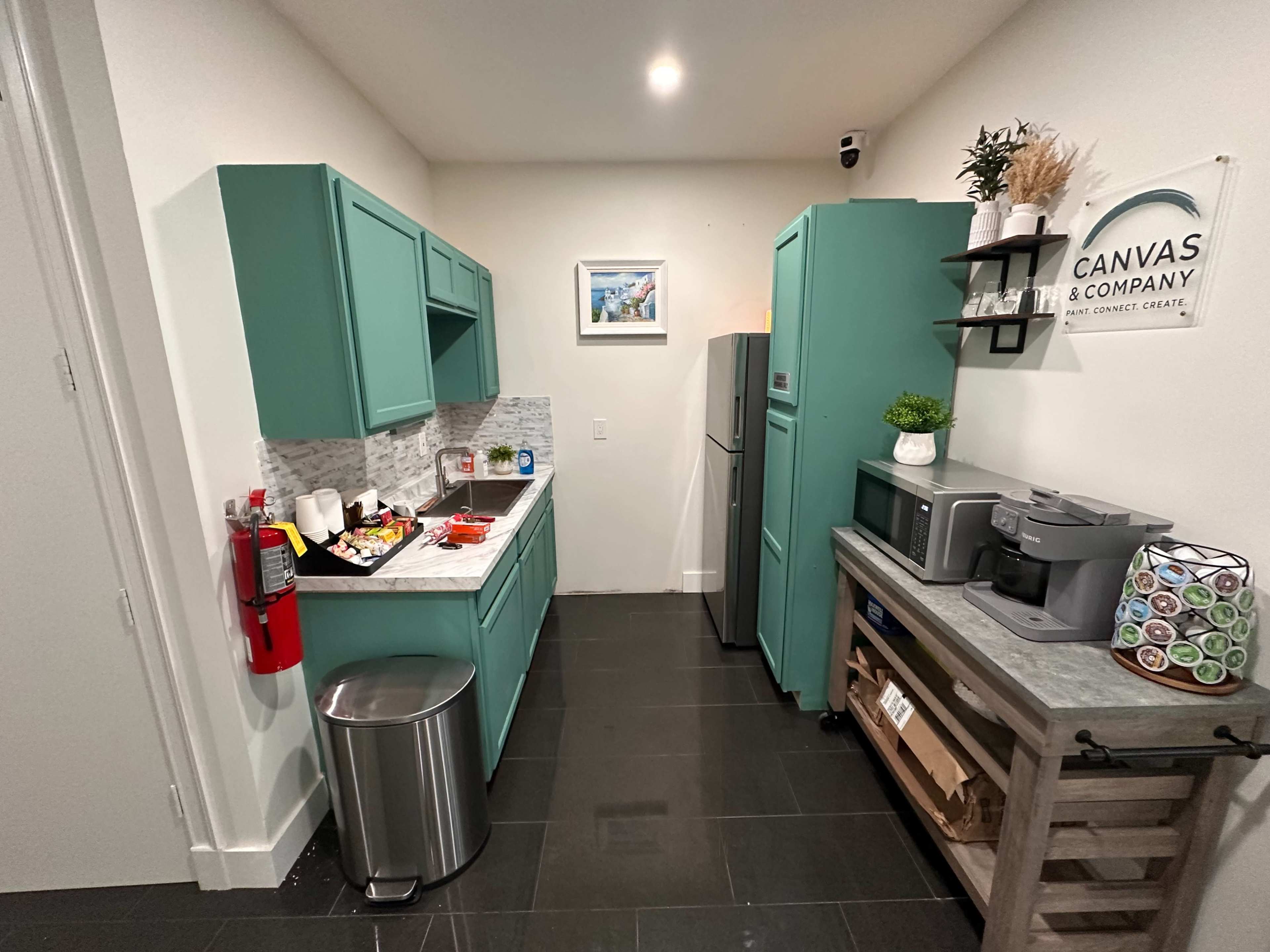 The image shows a small kitchen area with turquoise cabinets, a stainless steel refrigerator, and a countertop with snacks and a coffee maker.