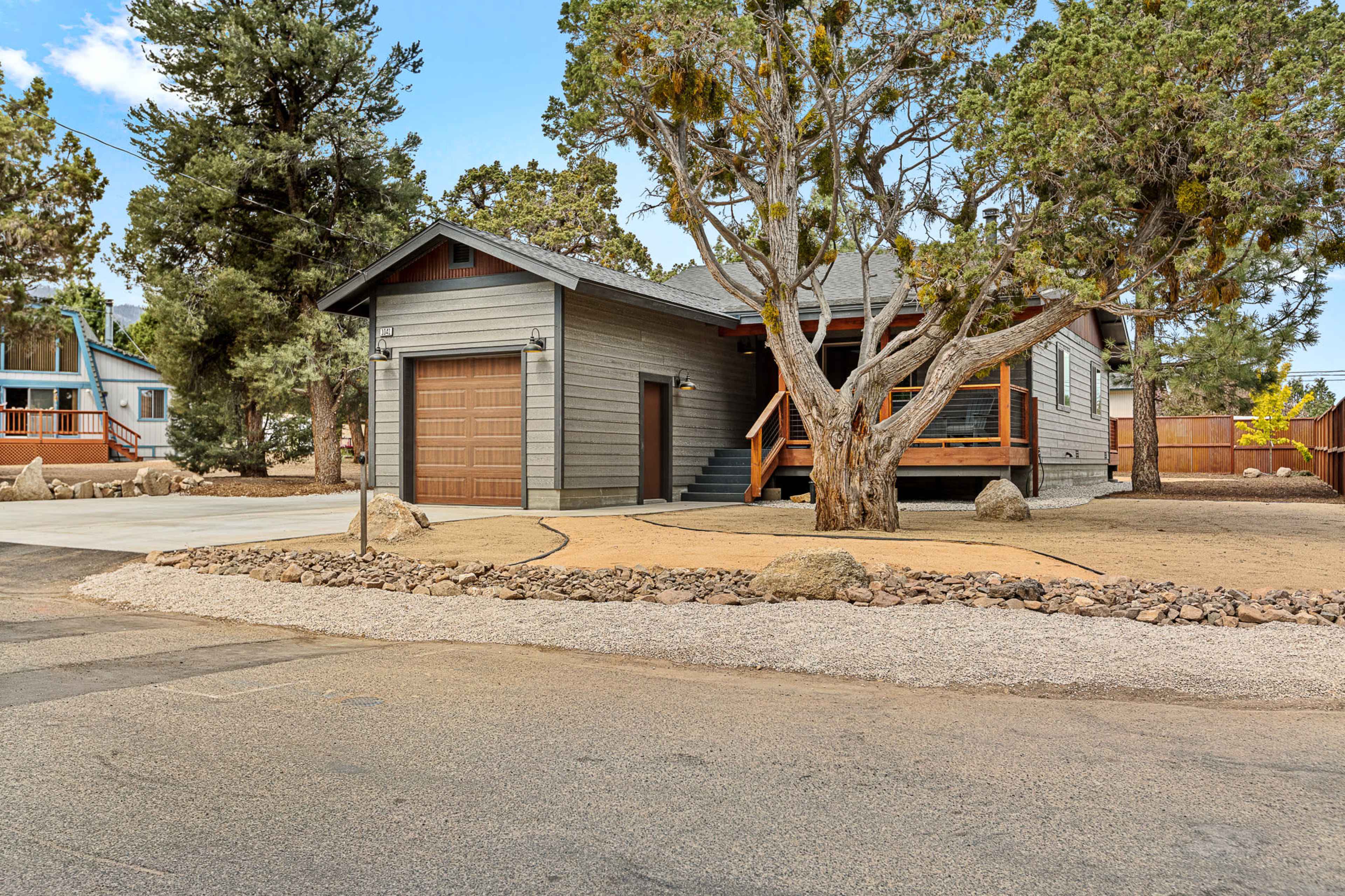 A single-story house with a garage is set on a gravel and dirt lot, surrounded by trees and rocky landscaping.