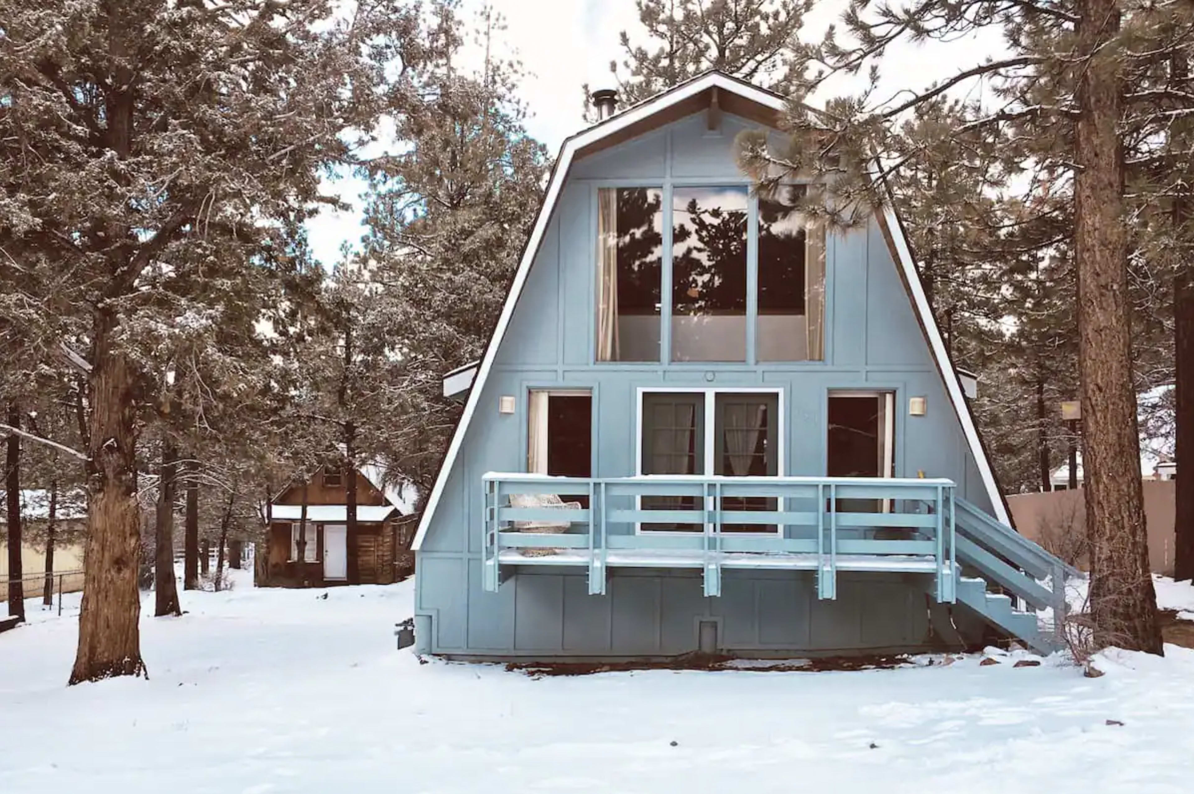 A blue A-frame cabin is surrounded by pine trees and covered in snow.