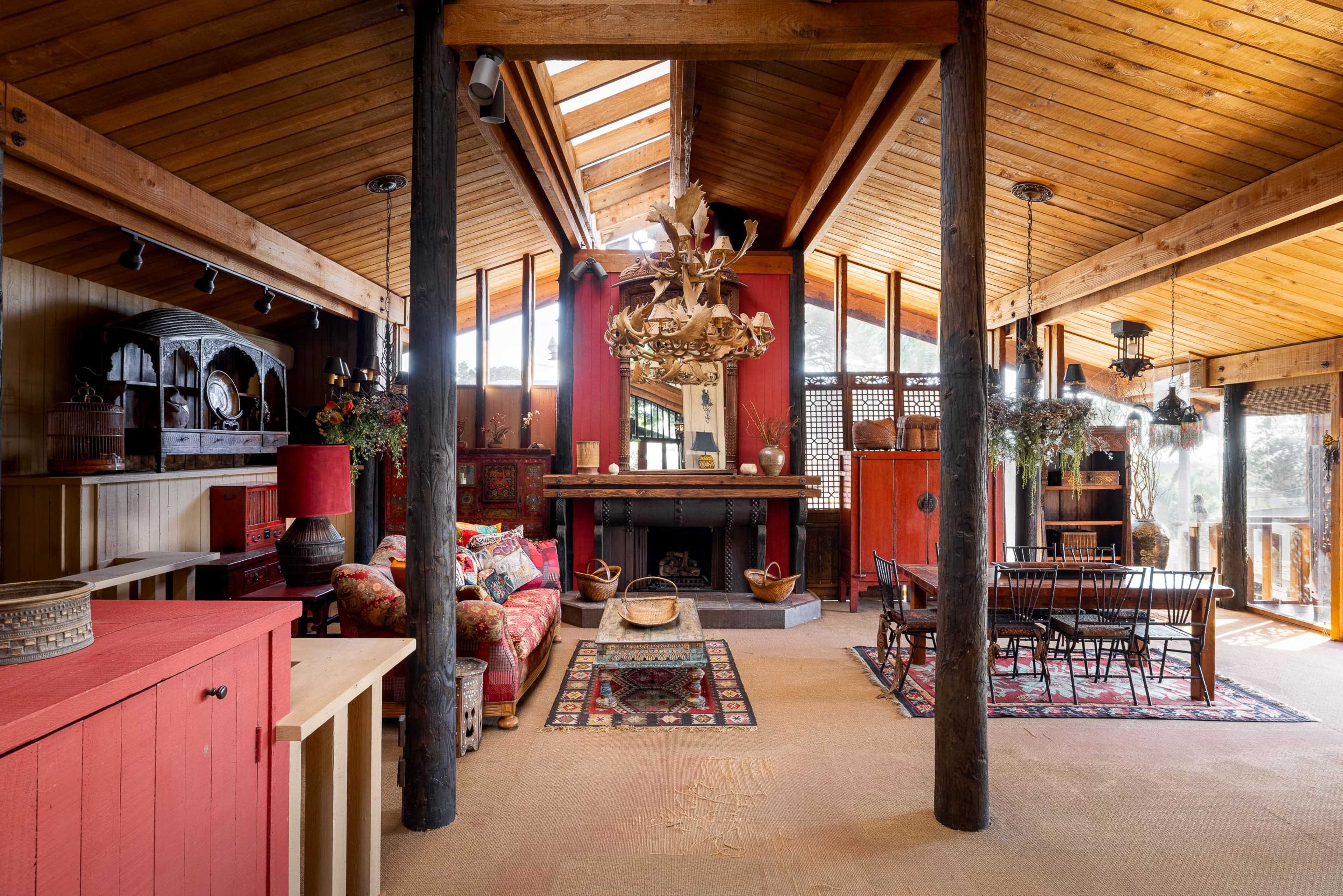 The image shows a spacious, rustic interior with wooden beams, a large chandelier, and a mix of furniture arranged around a central fireplace.