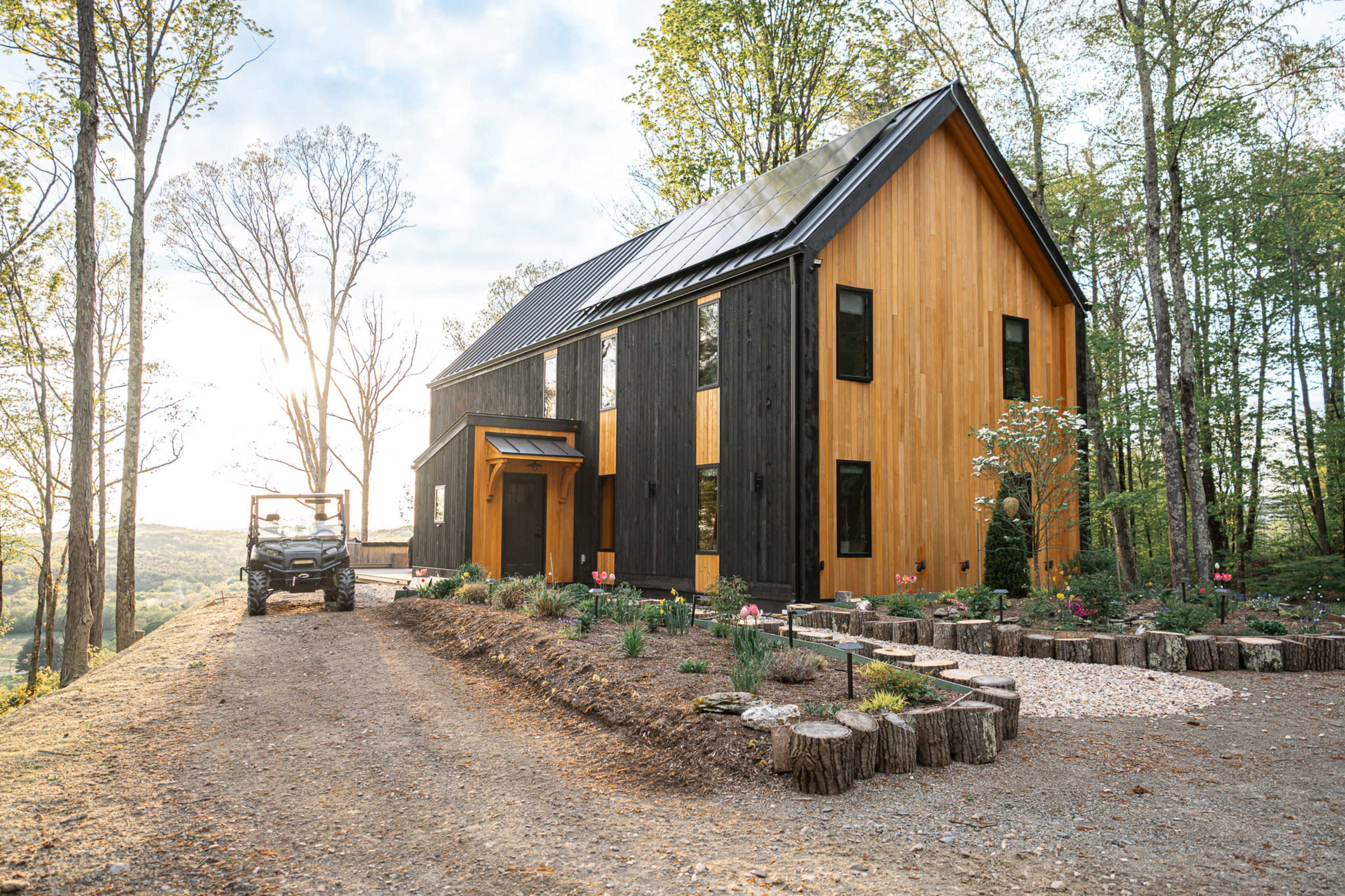 A modern wooden house with a black and orange exterior is situated on a dirt driveway surrounded by trees and landscaping.