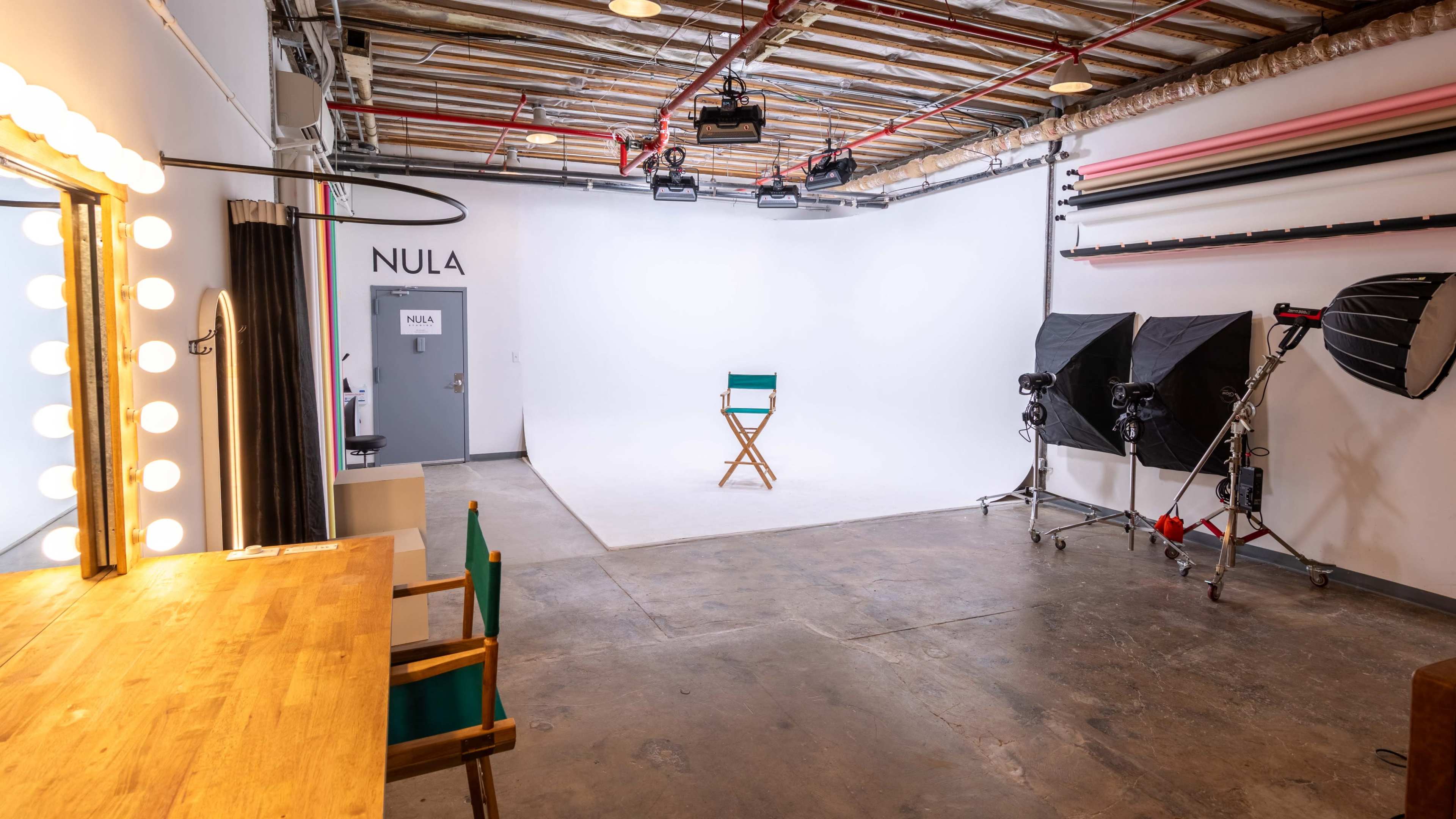 A minimalistic photography studio featuring a makeup area with a mirror, a single director's chair, and lighting equipment arranged around a plain white backdrop.