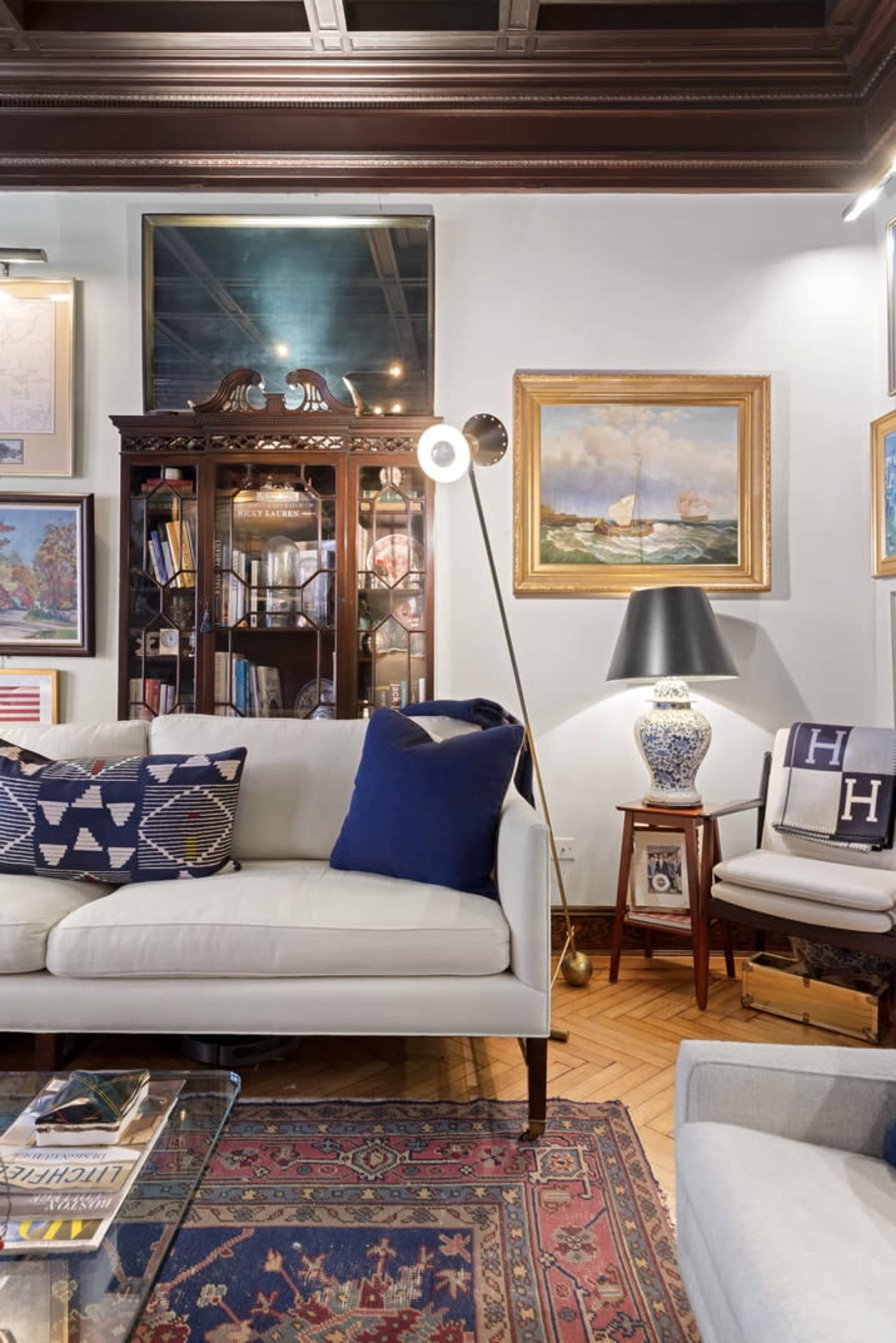 A cozy living room with a beige sofa, decorative pillows, a wooden cabinet displaying artwork, and a floor lamp beside a chair.