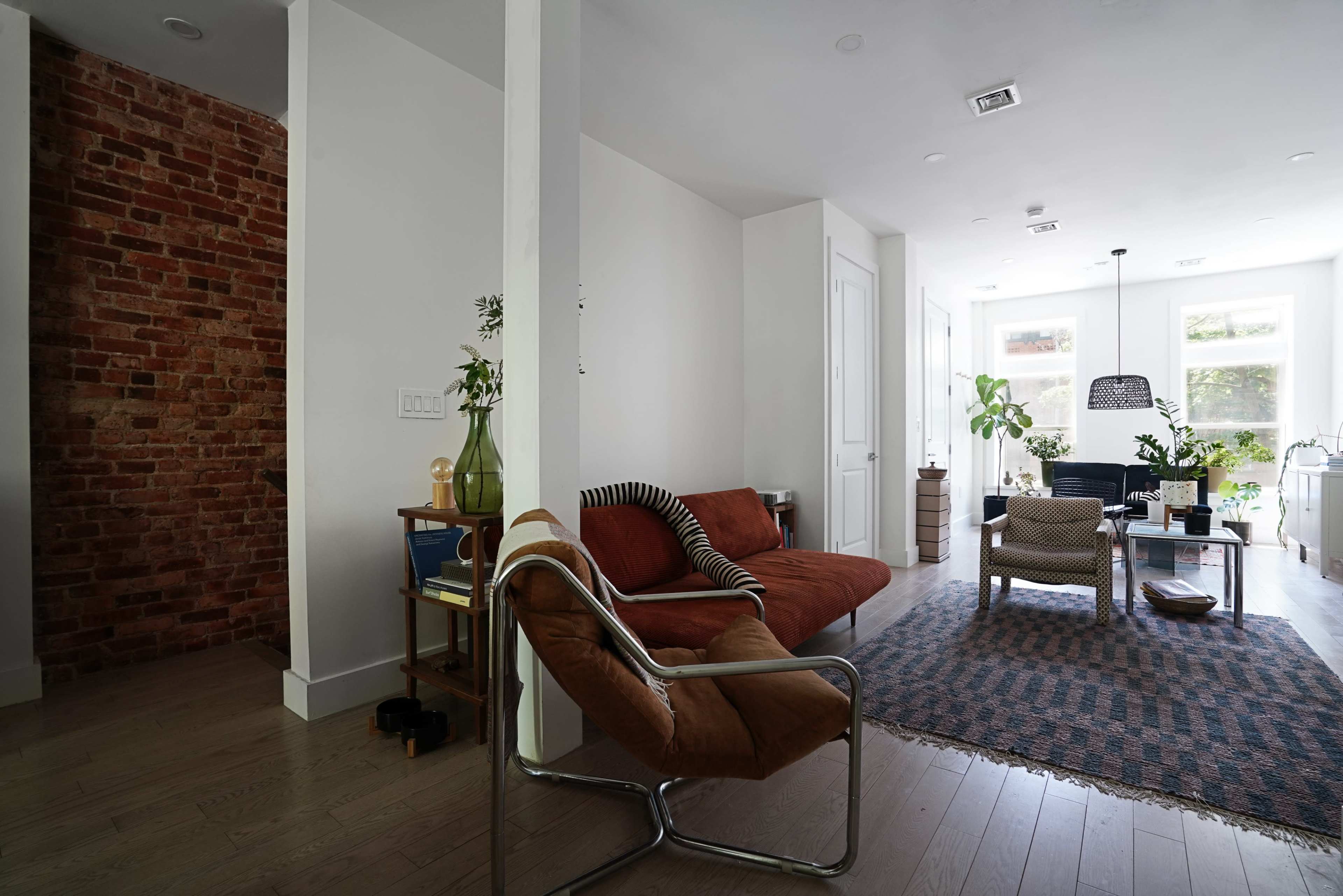 The image shows a modern living space featuring a brick accent wall, a brown sofa, a coffee table surrounded by chairs, and large windows bringing in natural light.