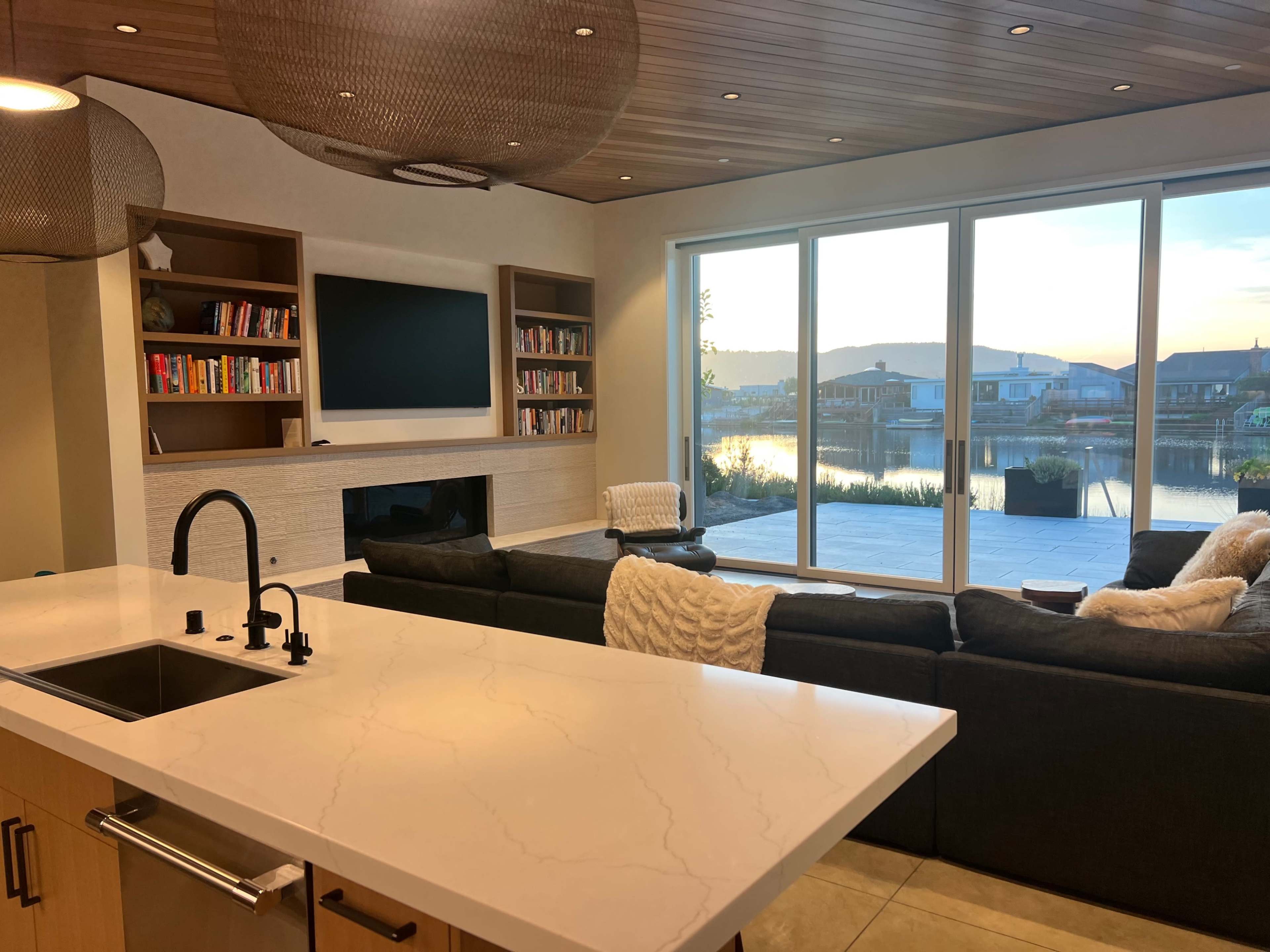 The image shows a modern living room with a large window overlooking a body of water, featuring a sectional sofa, a mounted television, and built-in bookshelves.