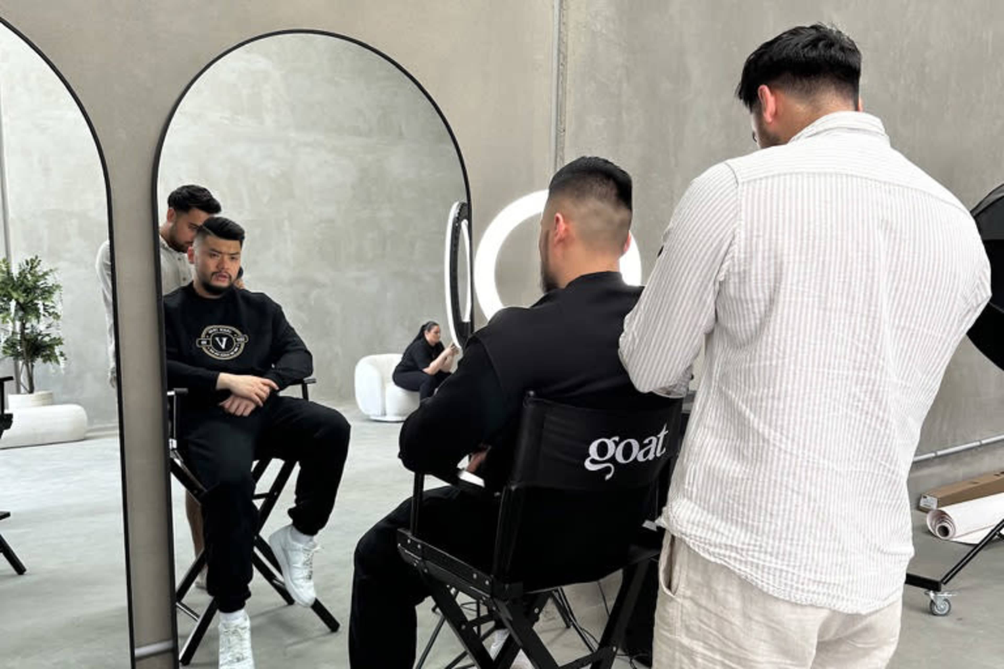 A man sits in a director's chair in front of a large mirror while another man prepares to style his hair.