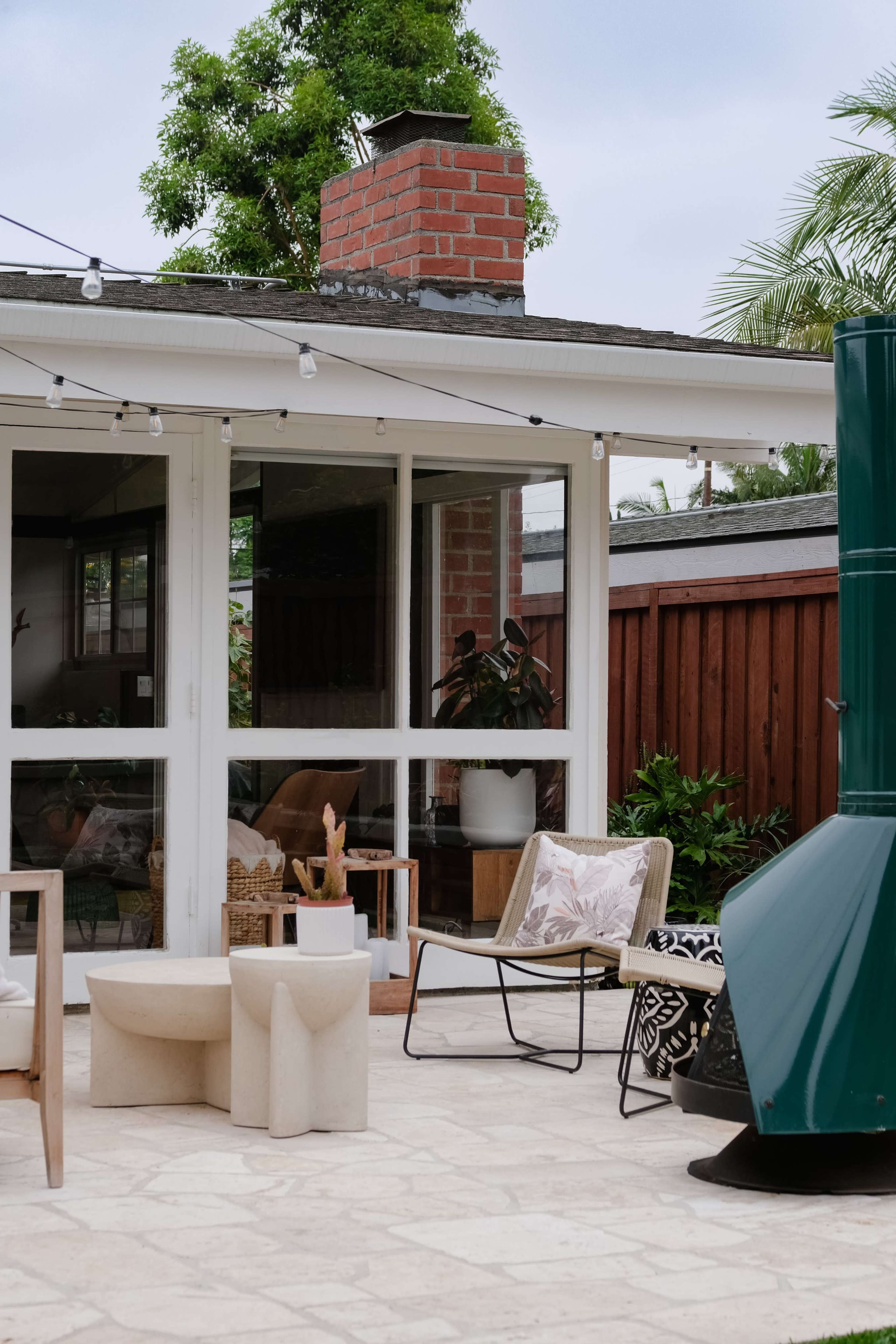 The image shows a patio area with modern outdoor furniture and decorative plants beside a brick chimney and a green fireplace.