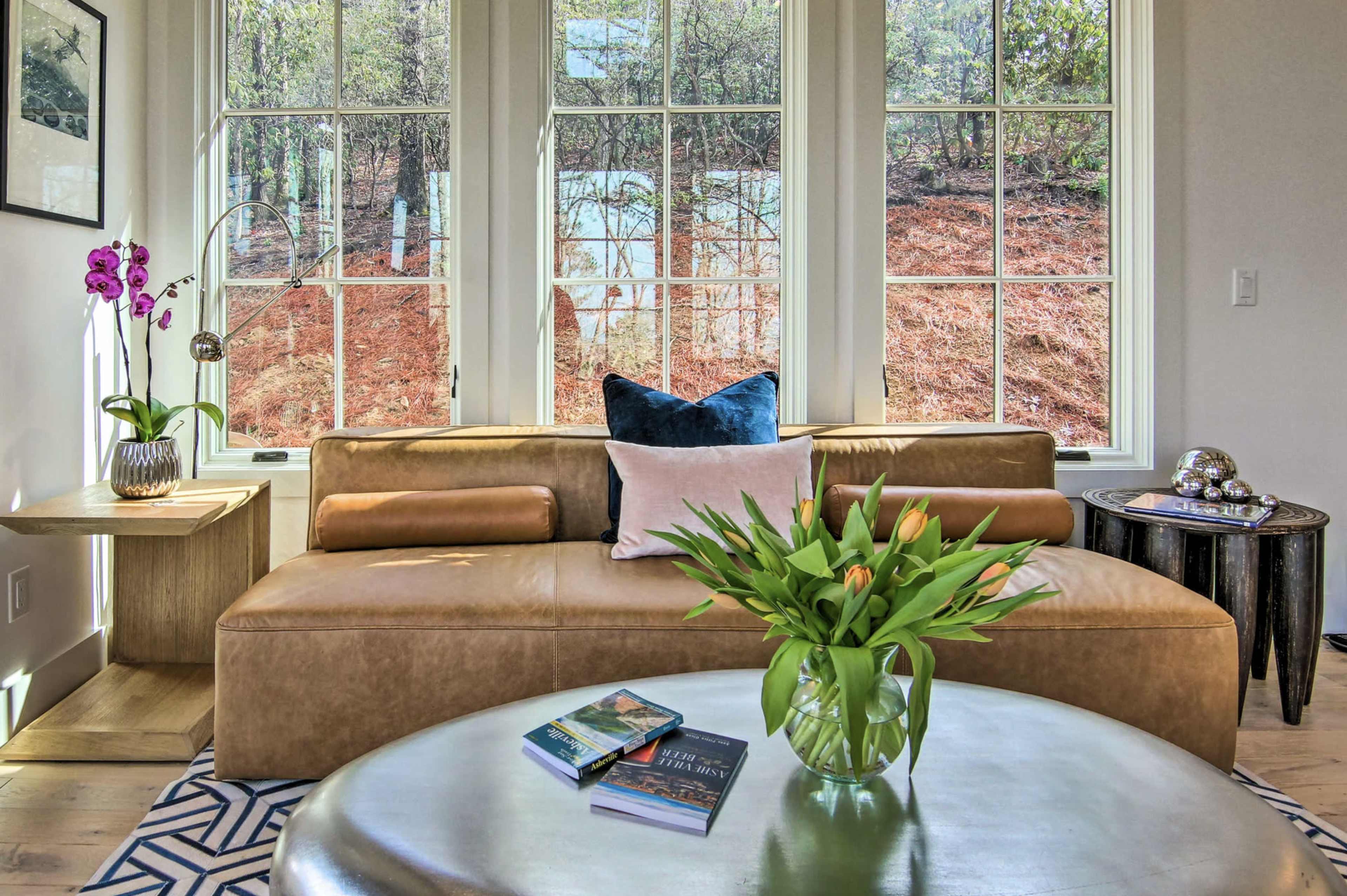 A modern living room features a tan leather sofa, round coffee table with books and flowers, and large windows overlooking a wooded area.