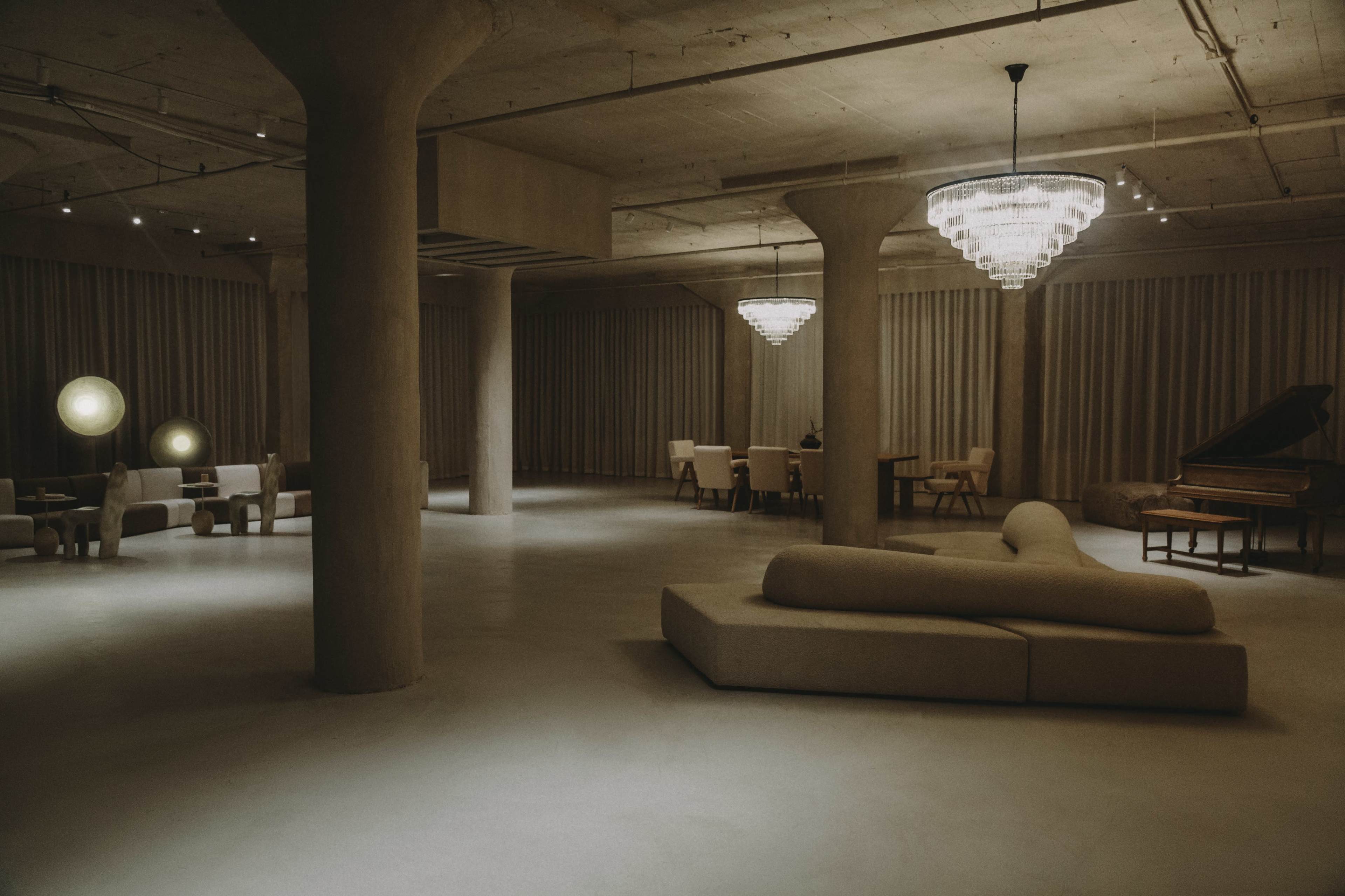 A spacious, dimly lit room with modern furniture, including a sectional sofa, dining area, and grand piano, surrounded by textured walls and soft drapery.