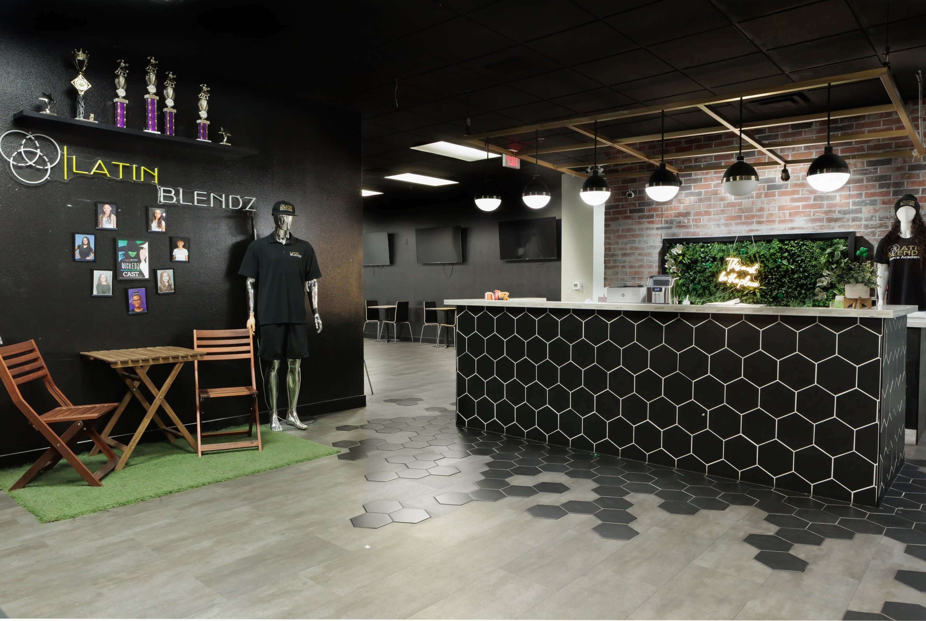 The image shows a modern café space with a hexagonal tile floor, a dark counter with a honeycomb pattern, and a seating area featuring a small wooden table and chairs.
