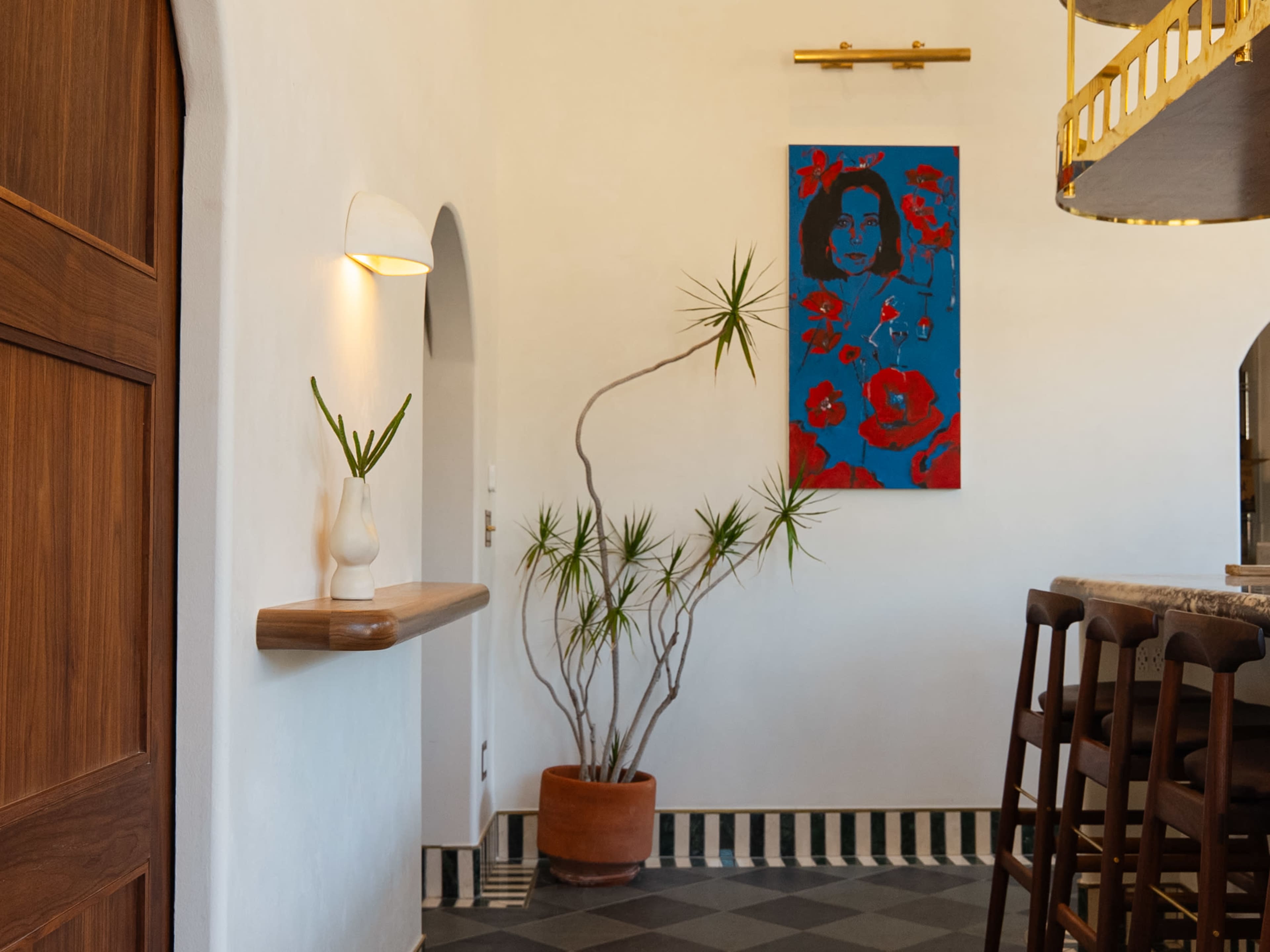 A modern interior features a wall with a colorful painting, a potted plant, and a wooden shelf, alongside a bar area with high stools.
