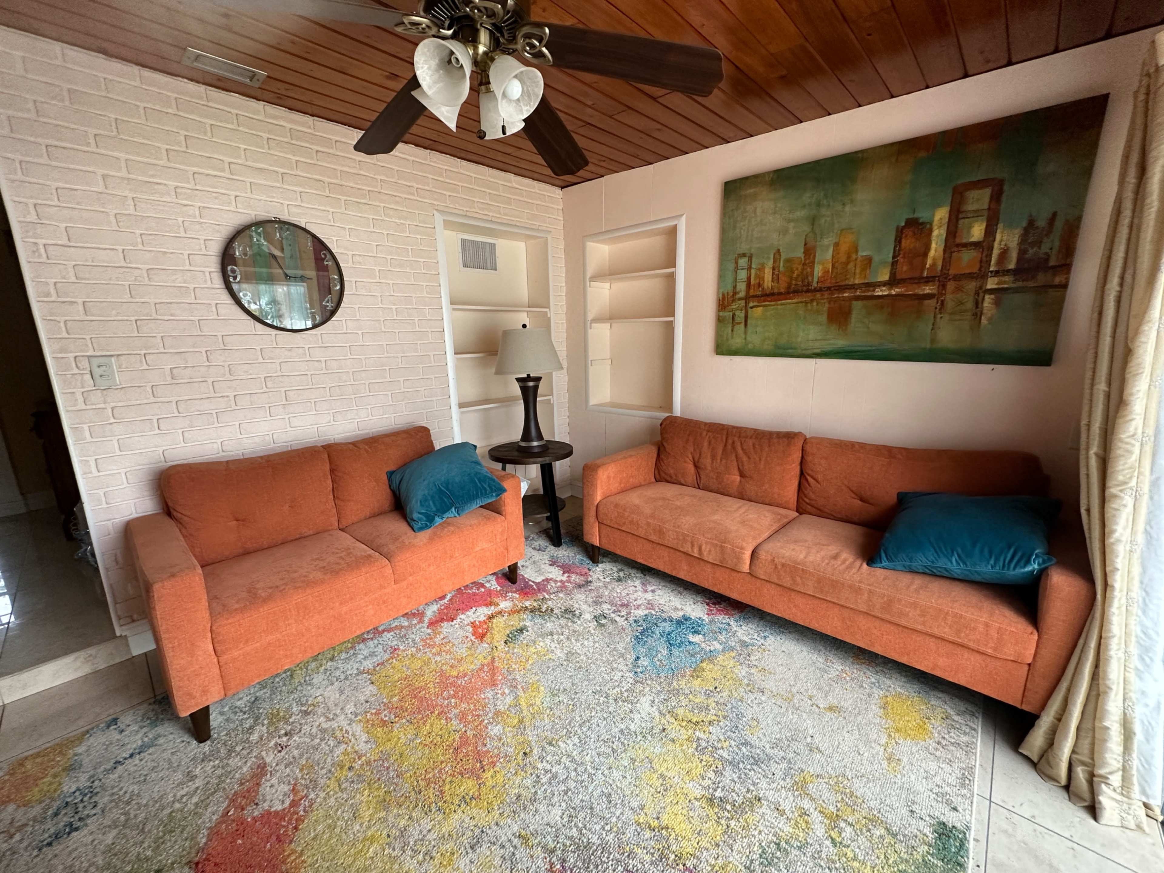 A living room with two orange sofas, a round mirror on the wall, a small table with a lamp, and a colorful area rug on the floor.