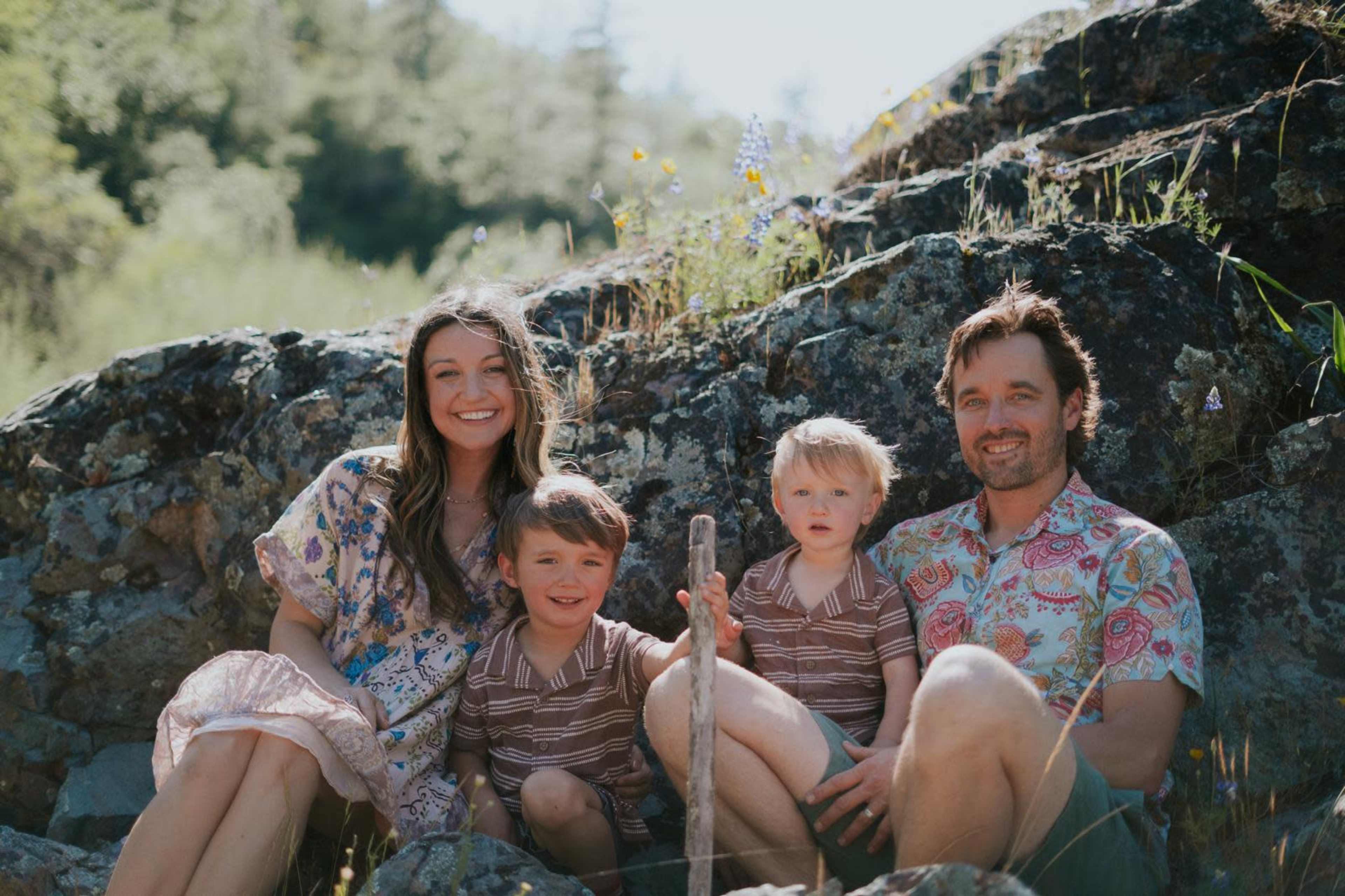 A family of four sits on a rocky hillside, smiling together in casual clothing amidst greenery and wildflowers.