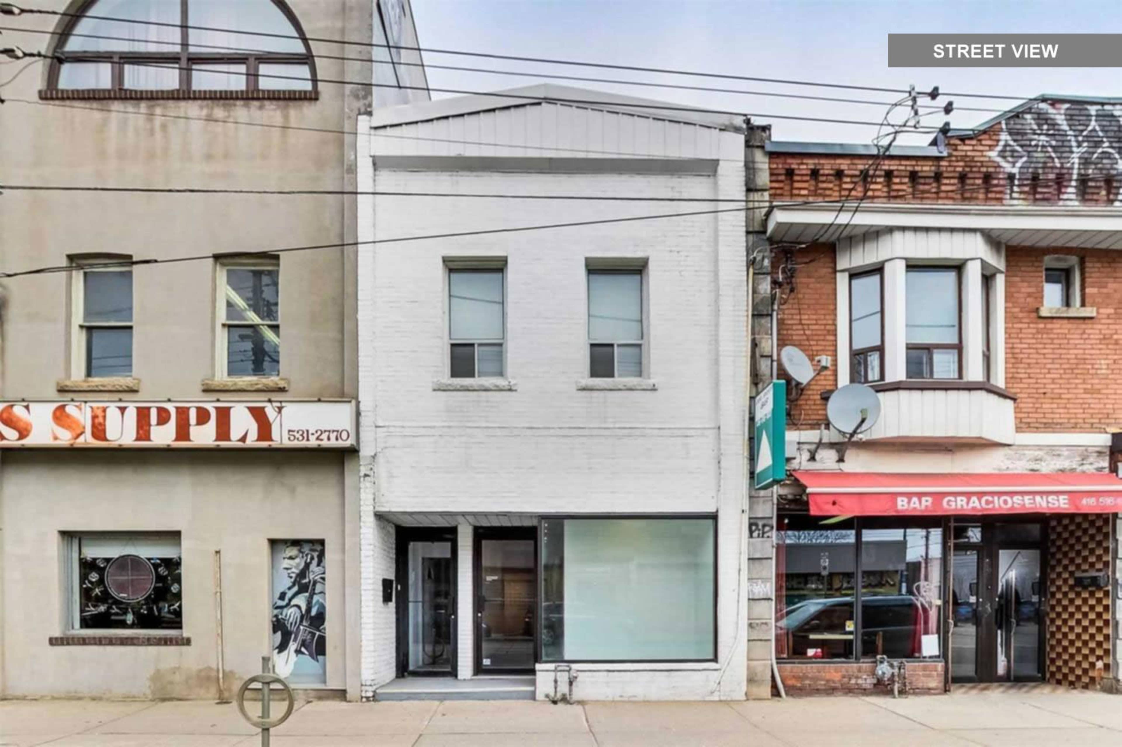 A two-story white building with large front windows, situated between a supply store and a bar on a city street.