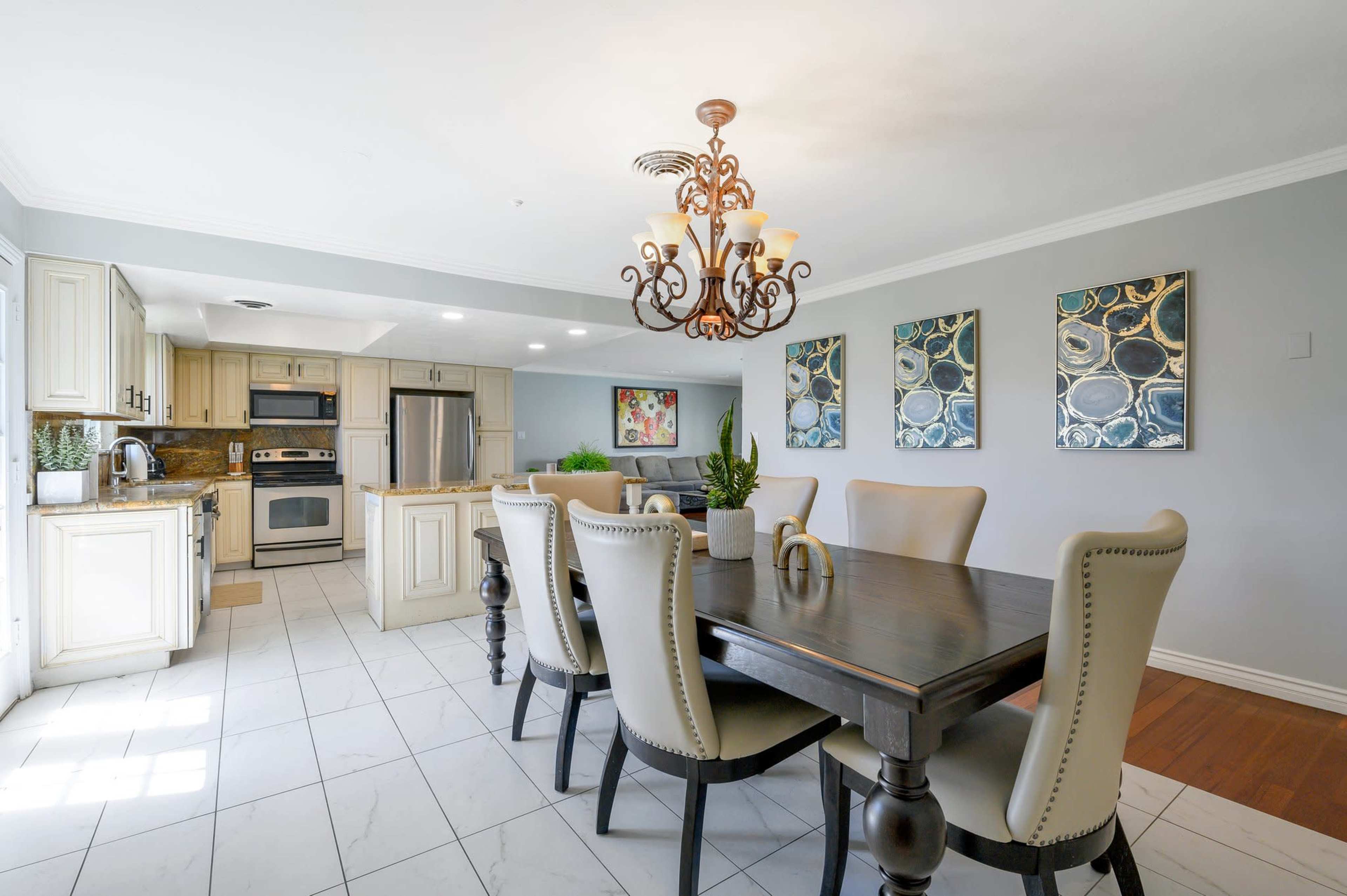 A modern kitchen and dining area featuring a wooden dining table with upholstered chairs, a chandelier, and stainless steel appliances.