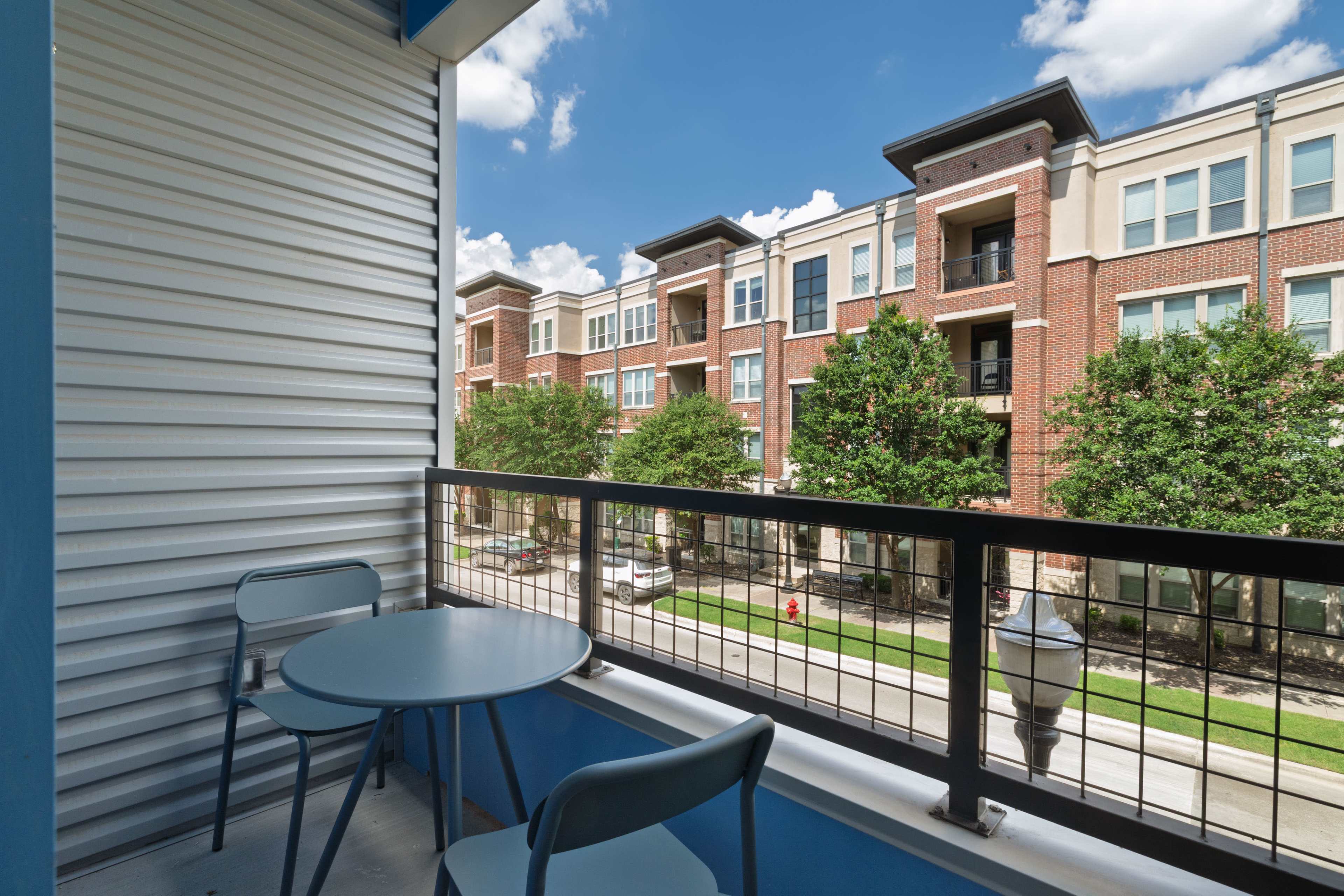 The image shows a balcony with a small round table and two chairs, overlooking a row of brick apartment buildings and greenery under a blue sky.