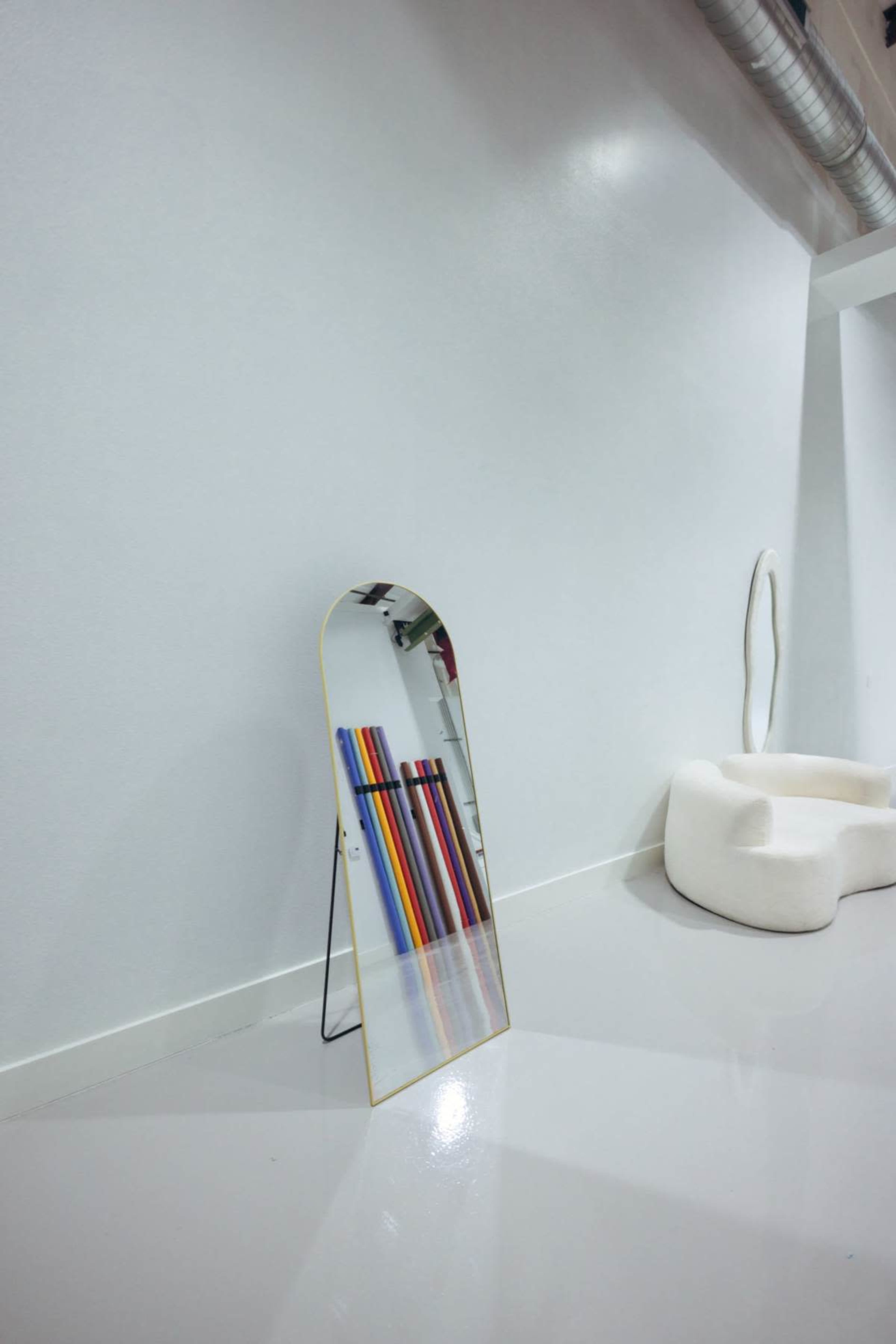 A tall, arched mirror stands on a black metal stand against a white wall, reflecting colorful vertical rods and a white, curved seating area nearby.