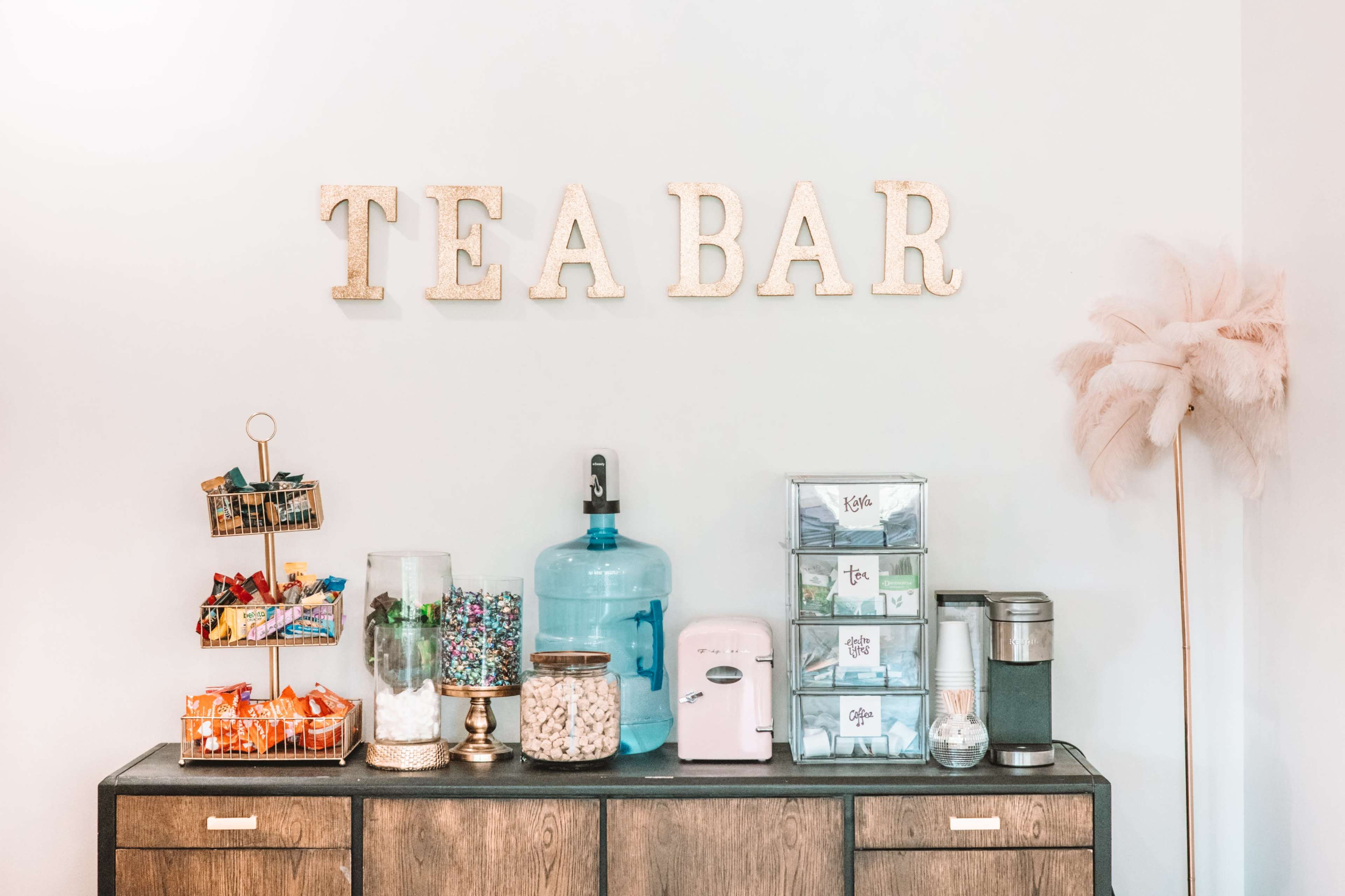 A tea bar features a variety of snacks and beverages, with a large water dispenser, a pink electric kettle, and decorative jars on a wooden surface.