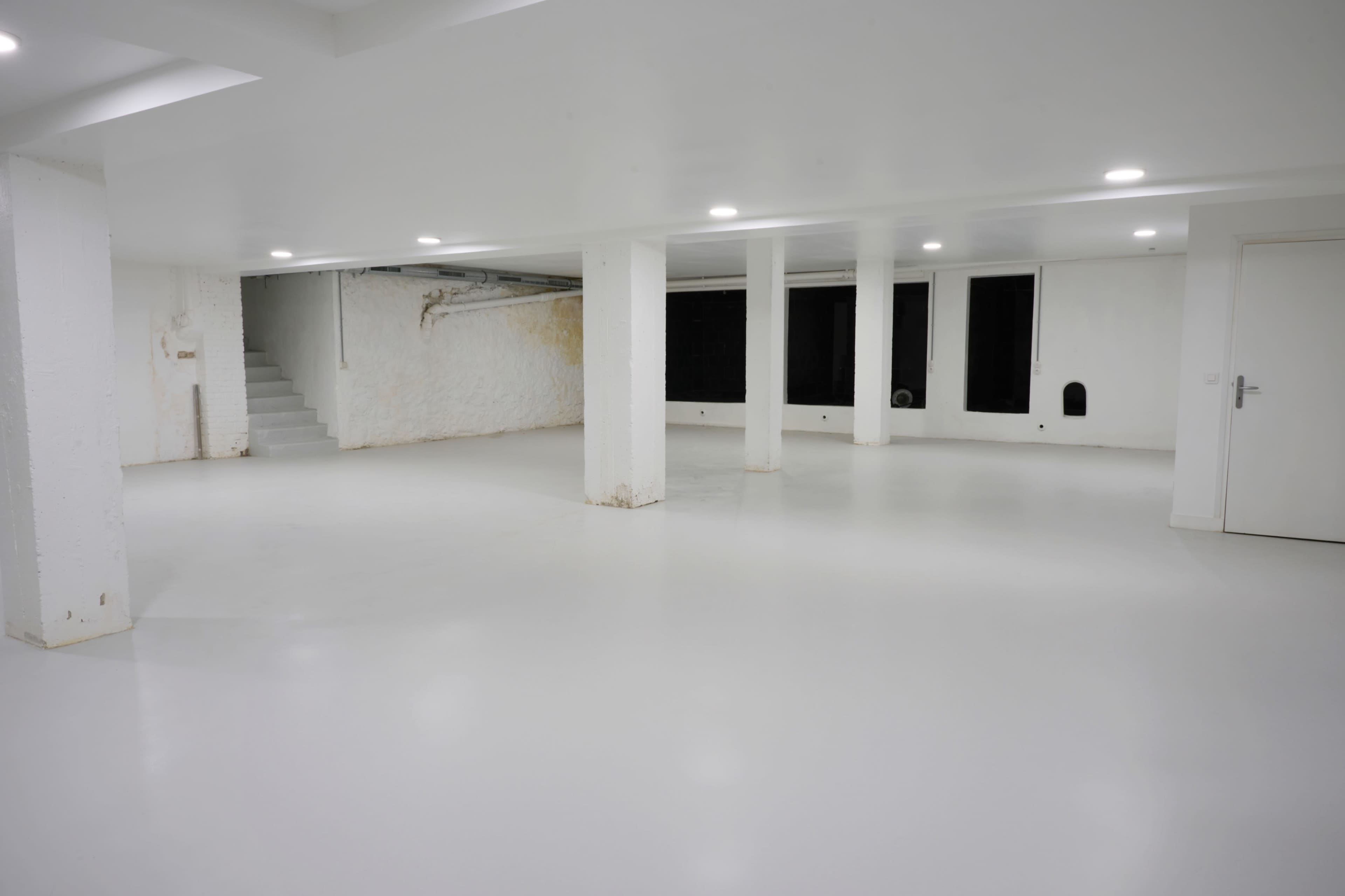 A spacious, empty room with a white floor, white walls, and pillars, illuminated by overhead lights.
