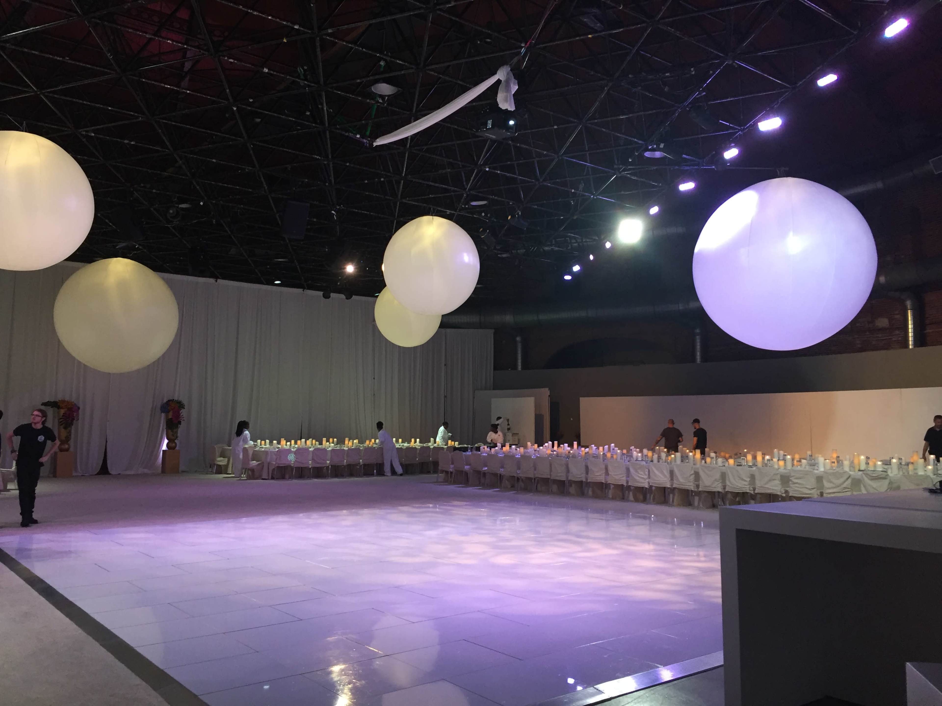 A spacious event venue set up for a formal gathering, featuring a long banquet table adorned with candles and large suspended round decorations.