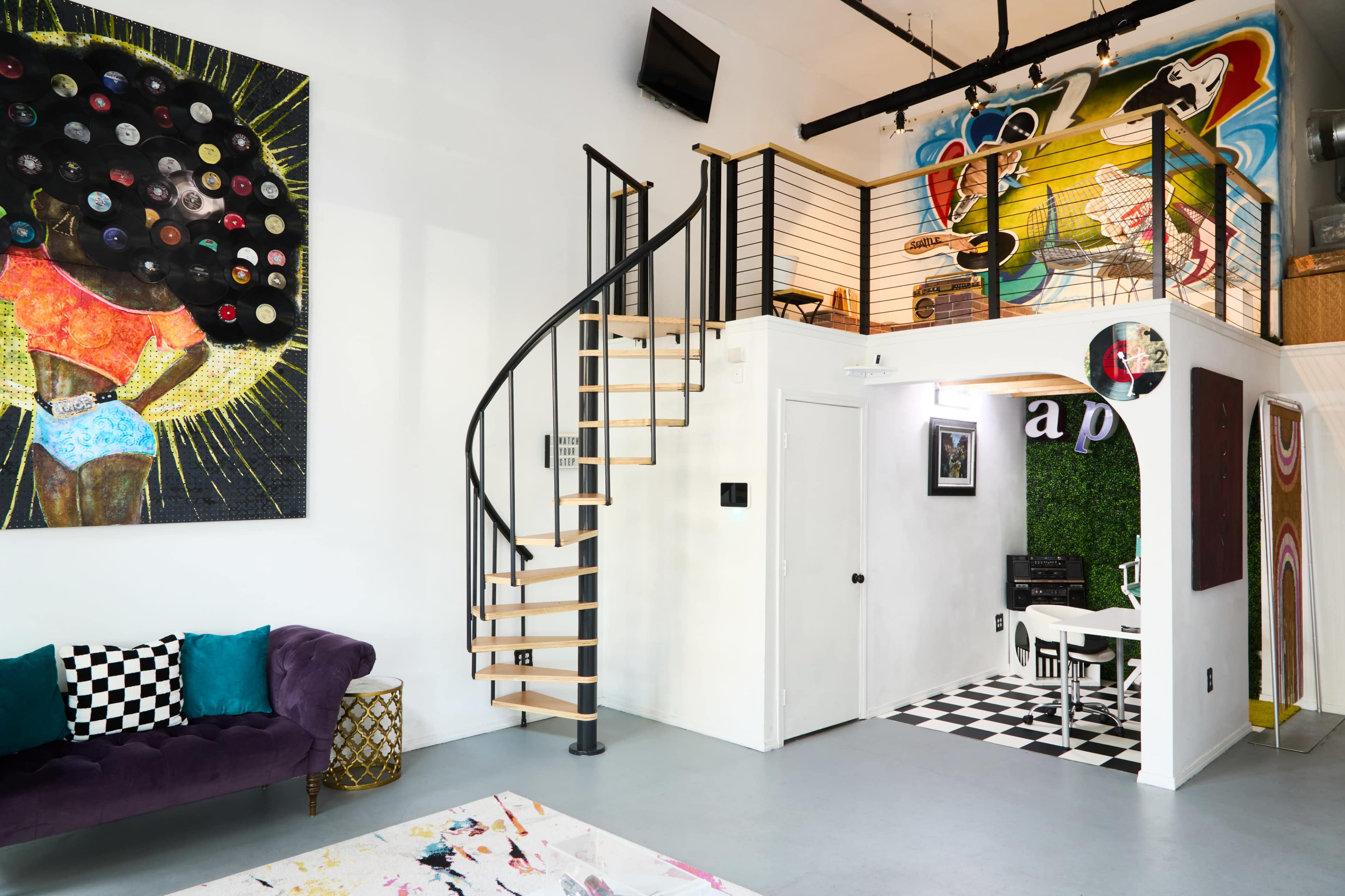 The image shows a modern interior featuring a spiral staircase, vibrant artwork on the walls, and a cozy seating area with a checkered pattern.