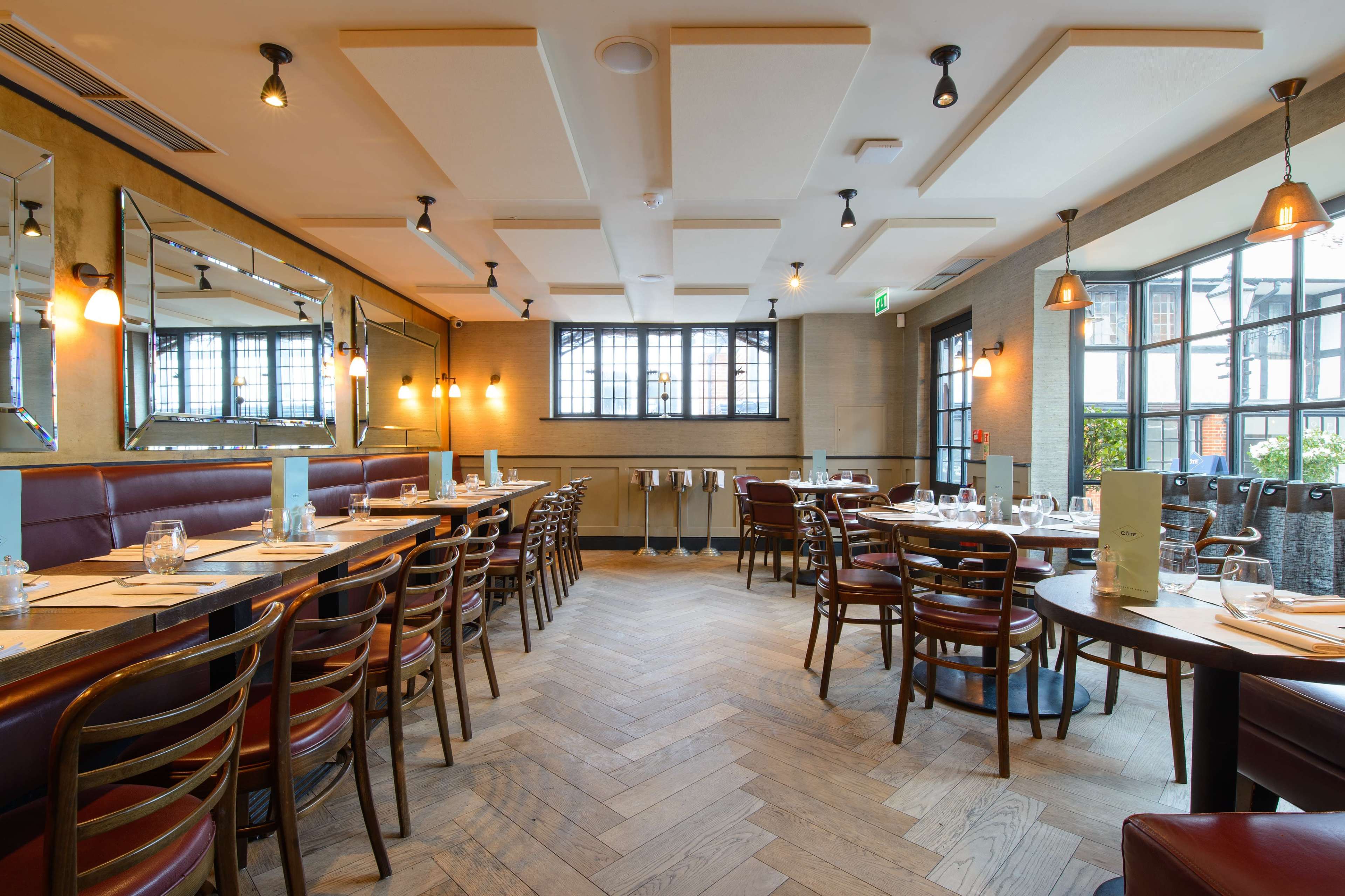 The image depicts a well-lit restaurant interior with wooden floors, a combination of round and rectangular tables, and mirrored walls.