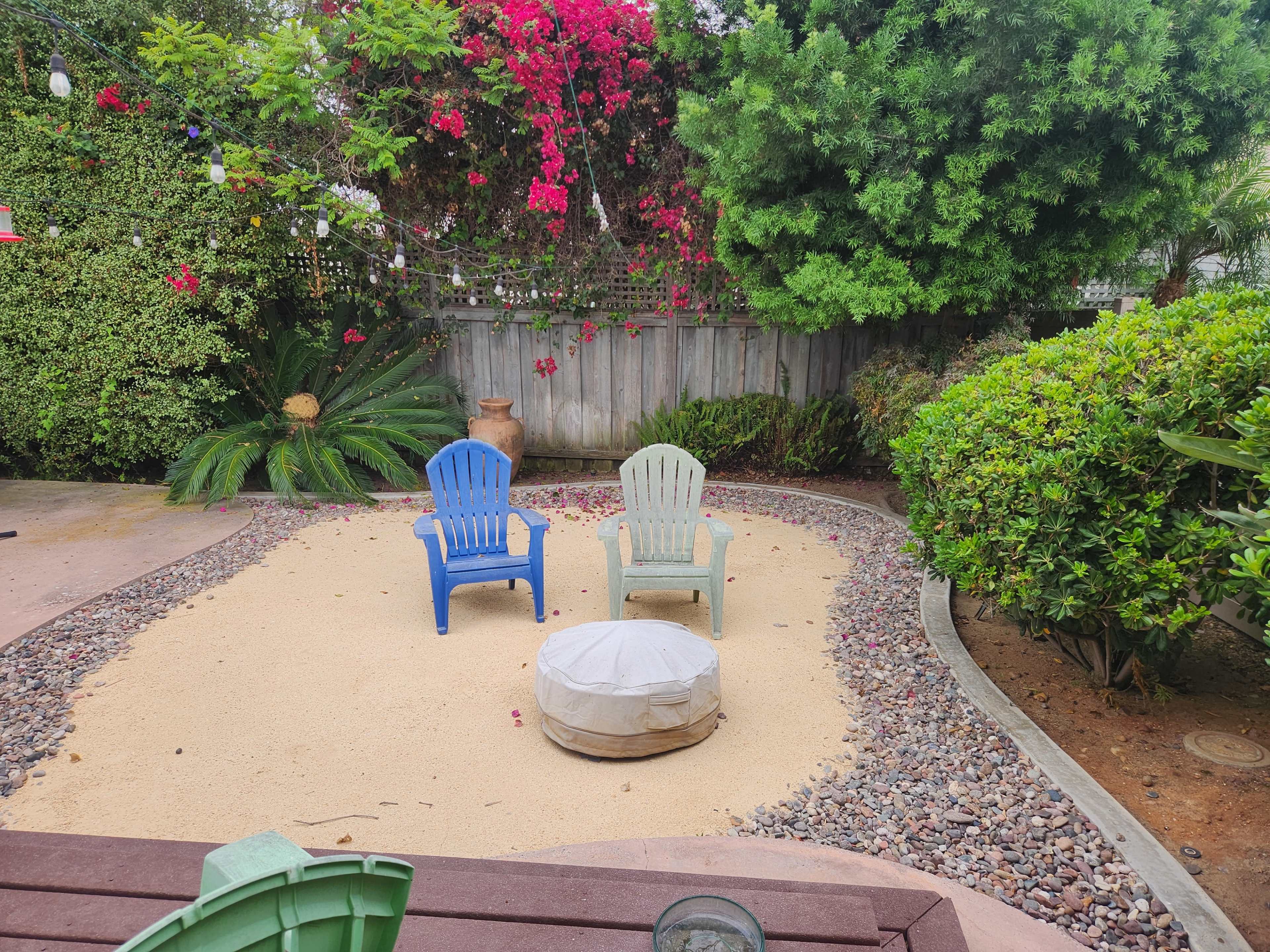 The image shows a sandy area in a garden with two plastic chairs and a round covered object surrounded by greenery and flowering plants.