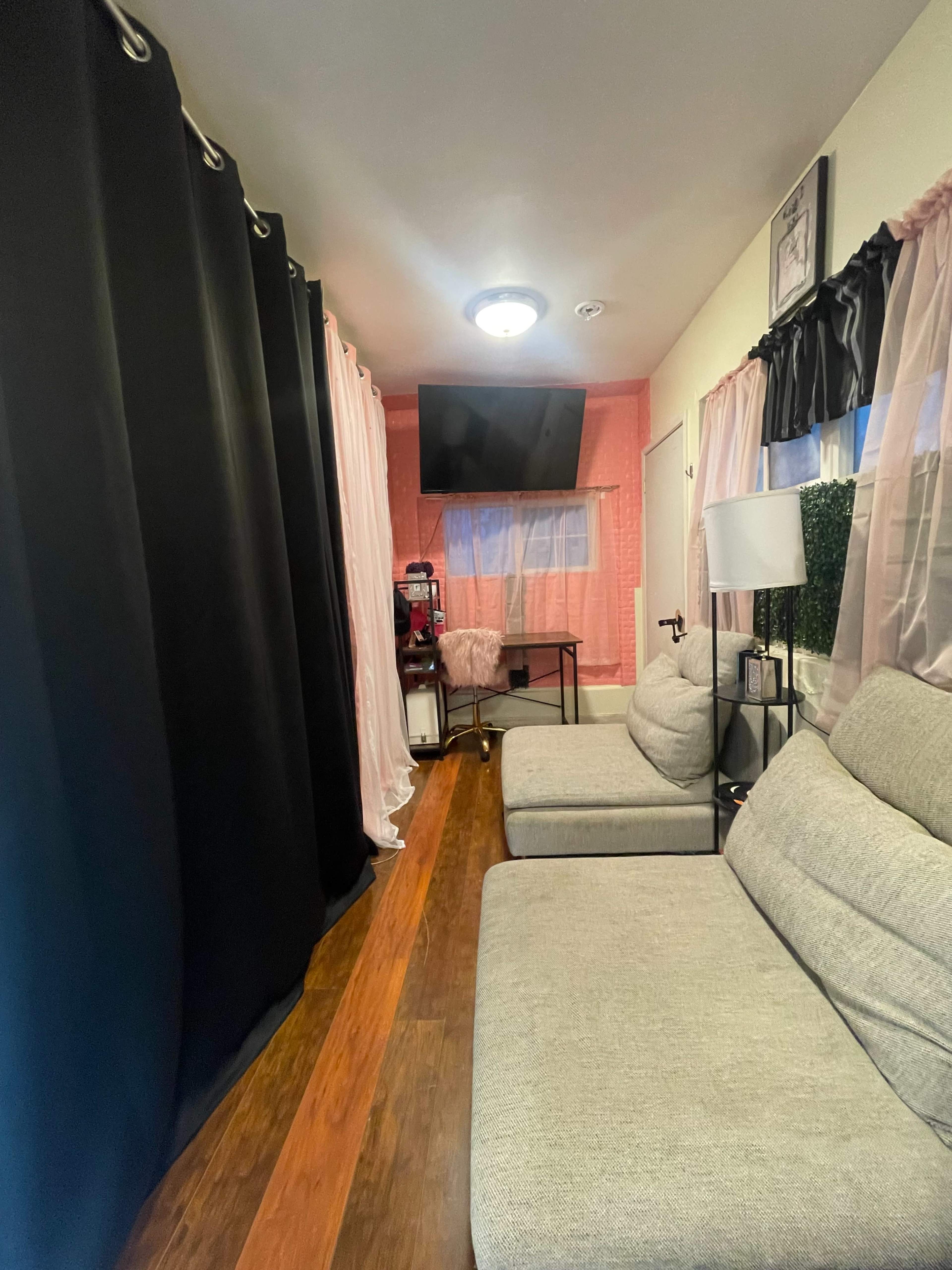 A narrow living space features two gray sofas facing a black television mounted on the wall, with curtains in black and pink separating the area.
