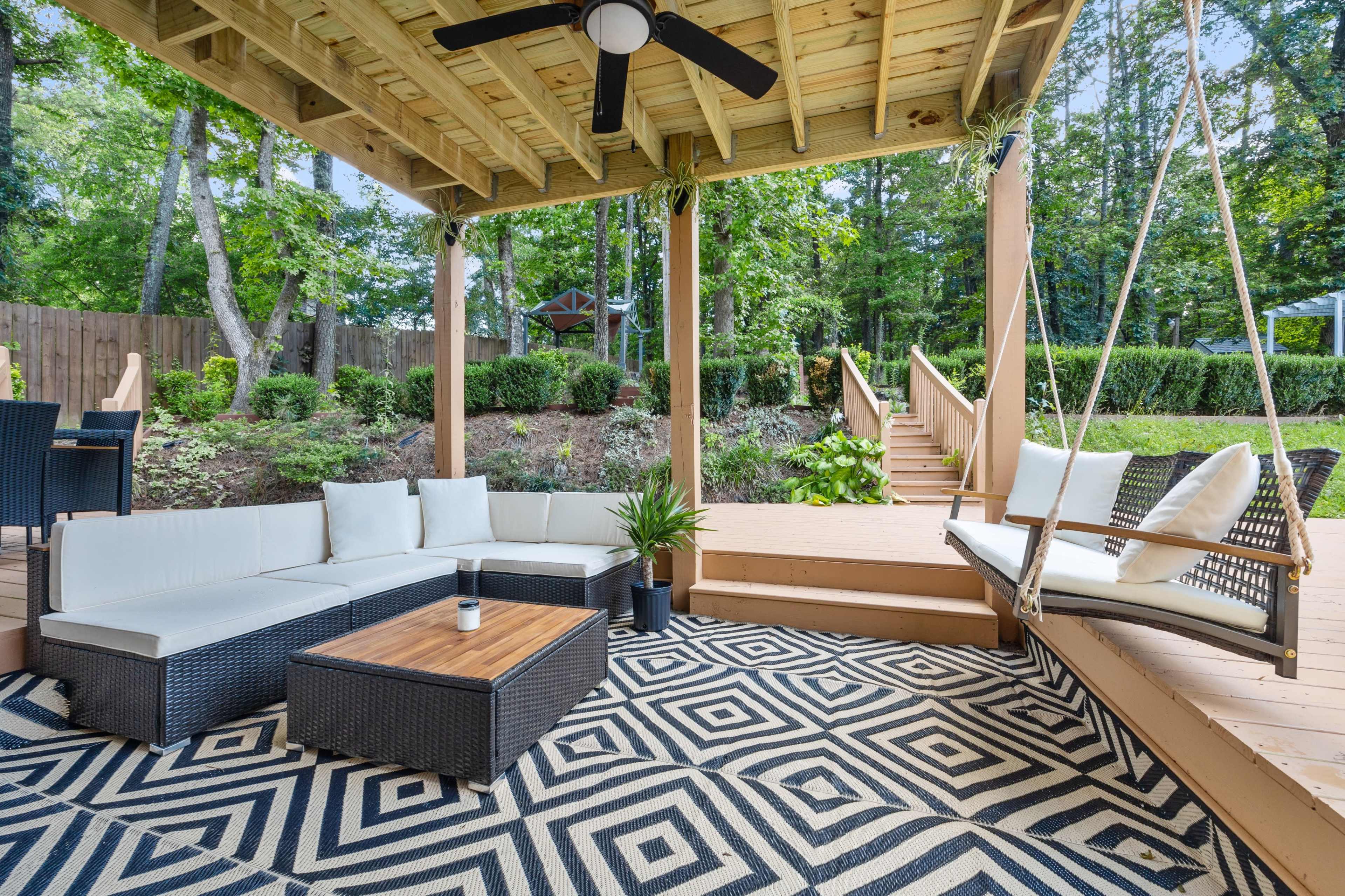 The image shows a covered outdoor seating area with a sectional sofa, a coffee table, and a swing, surrounded by greenery and leading to a wooden staircase.