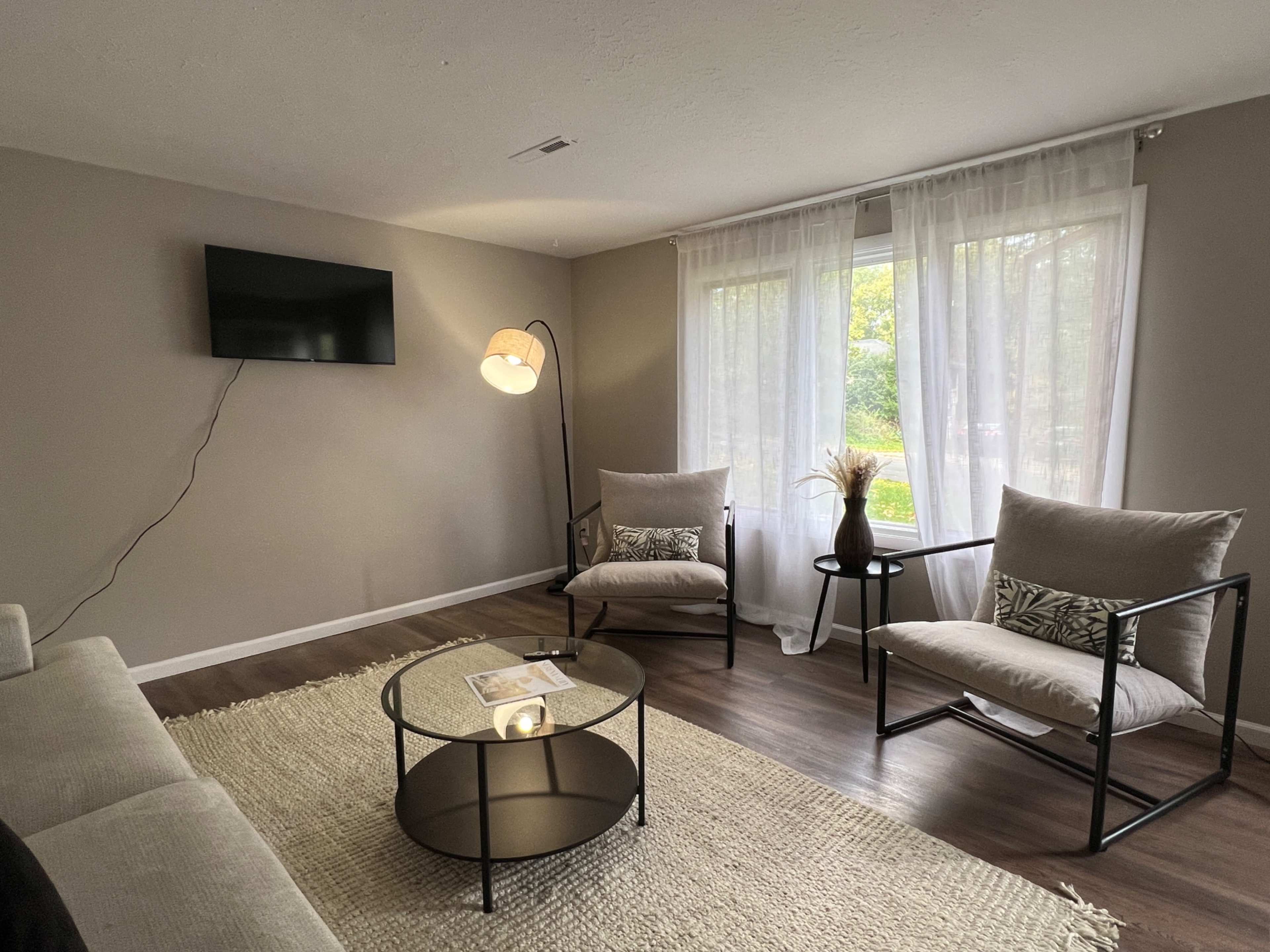 A cozy living room features a television mounted on the wall, two armchairs, a round coffee table, and large windows with sheer curtains allowing natural light.
