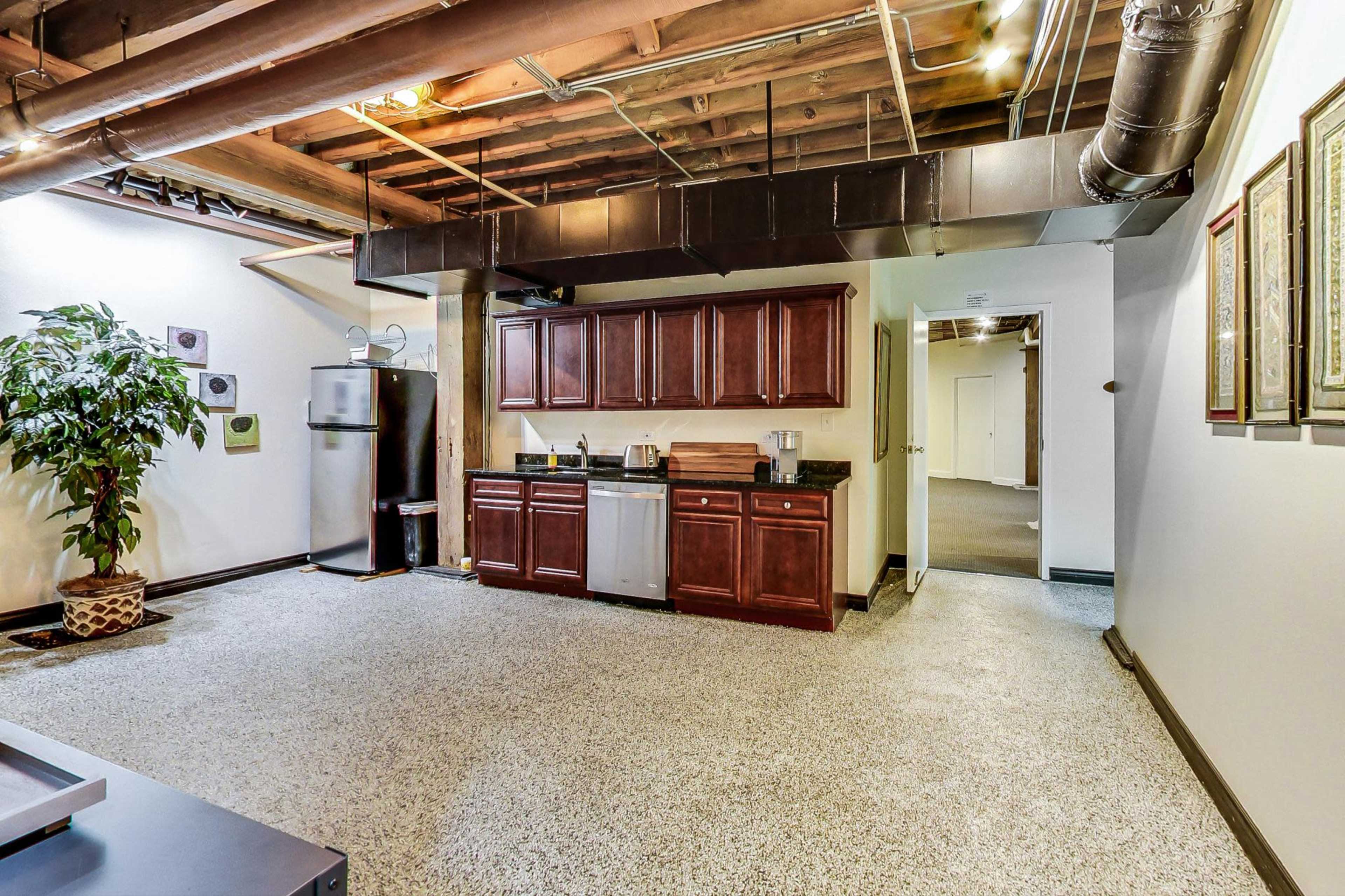 A basement area with a kitchenette featuring wooden cabinets, a countertop, a small refrigerator, and a decorative plant in the corner.