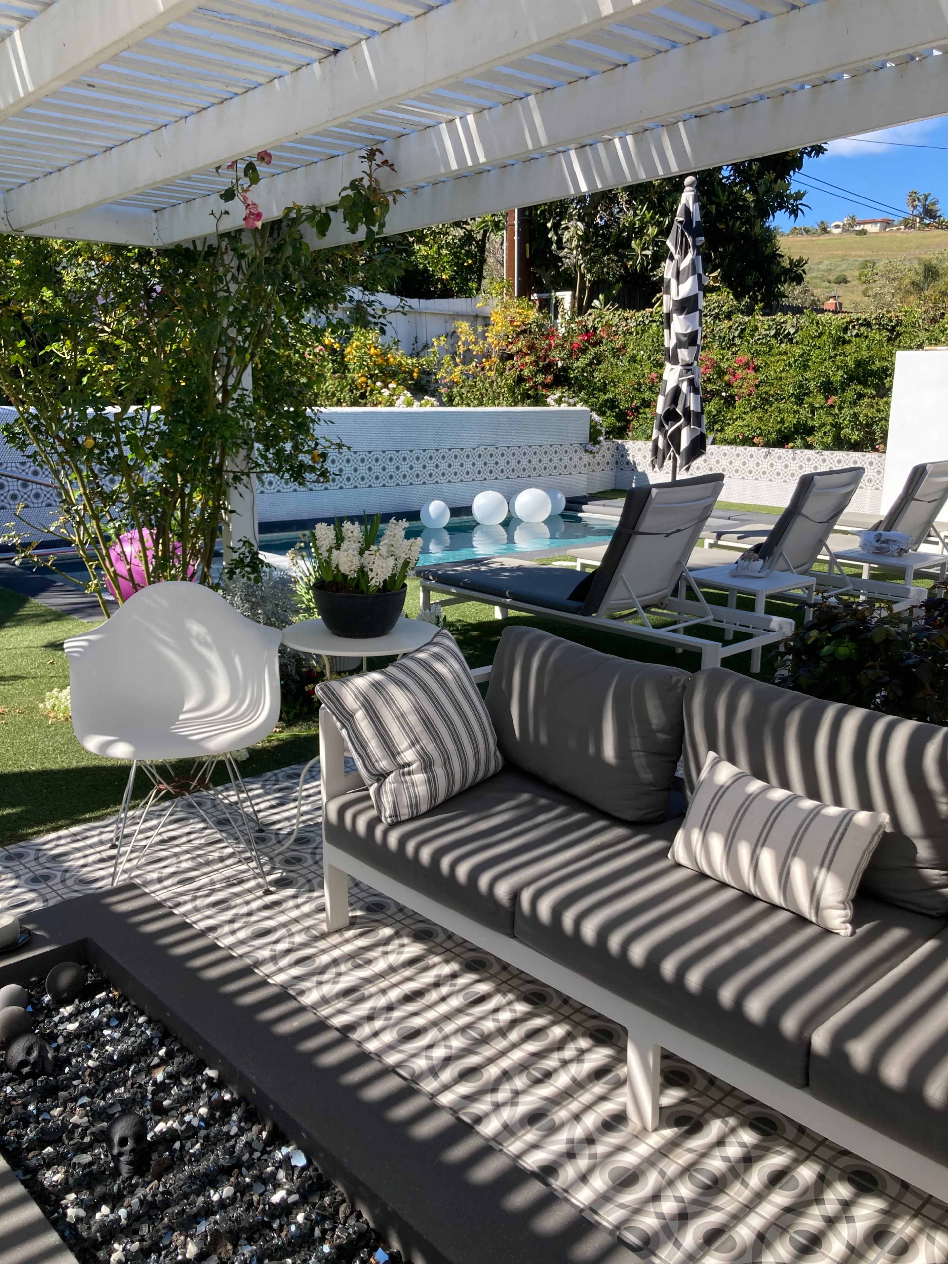 A modern outdoor seating area with a gray sofa, decorative pillows, and a view of a pool surrounded by lounge chairs and trees under a pergola with striped shadows.
