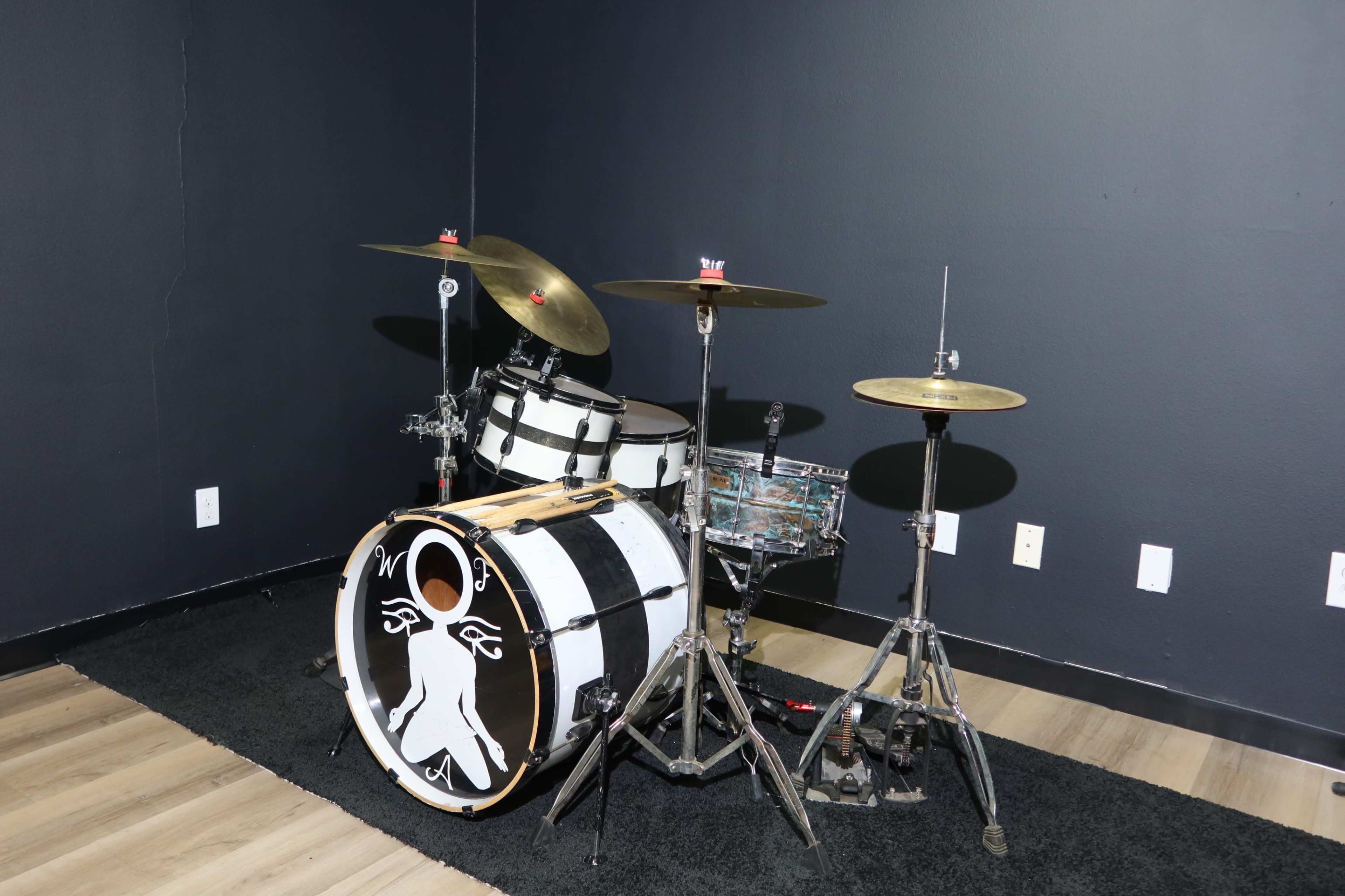 The image shows a drum kit set up against a dark wall, featuring a large bass drum with a graphic design and two cymbals mounted on stands.