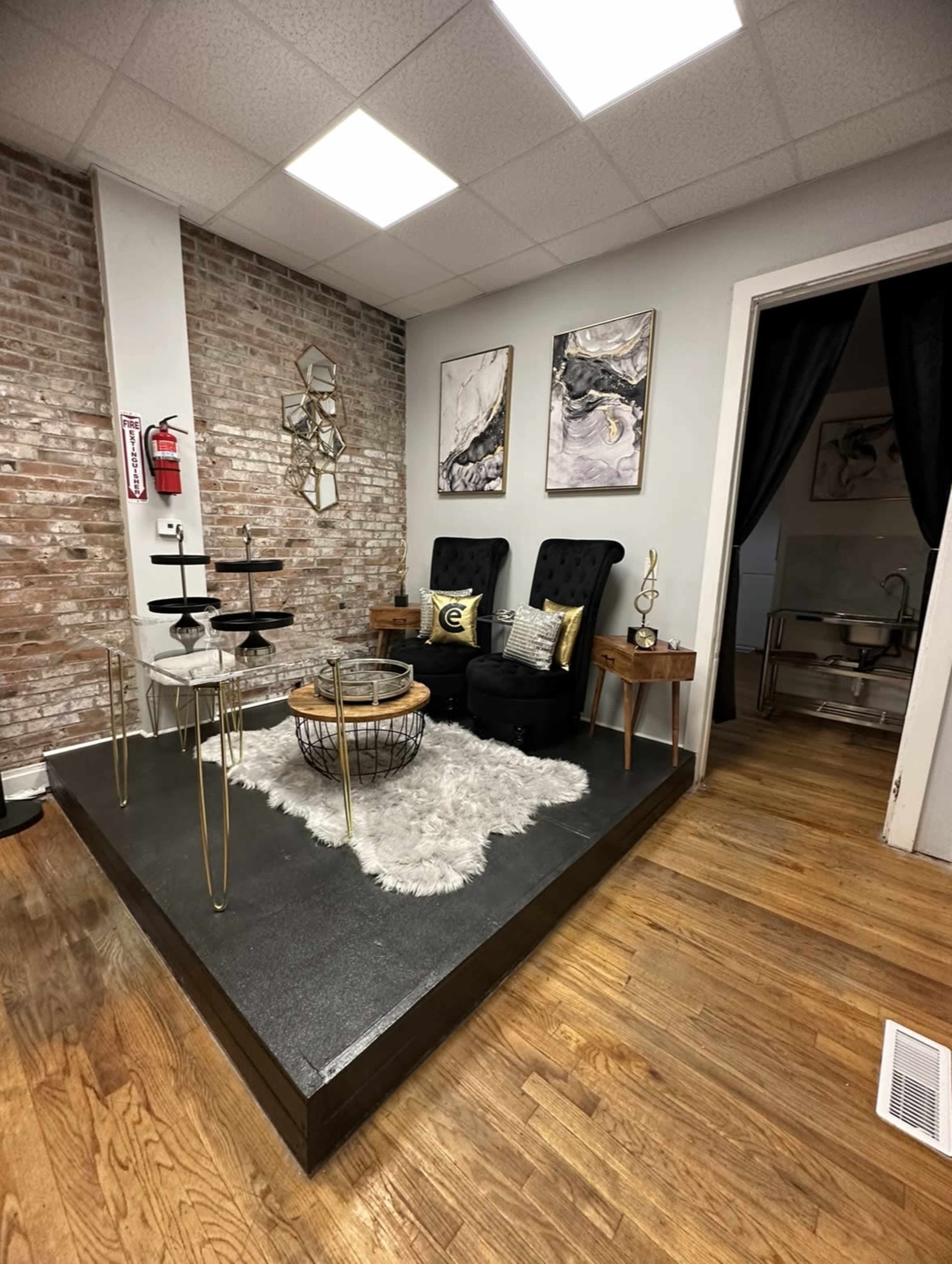 The image shows a cozy corner of a room featuring two black chairs on a raised platform, a round table, decorative items, and a partially visible area behind a curtain.