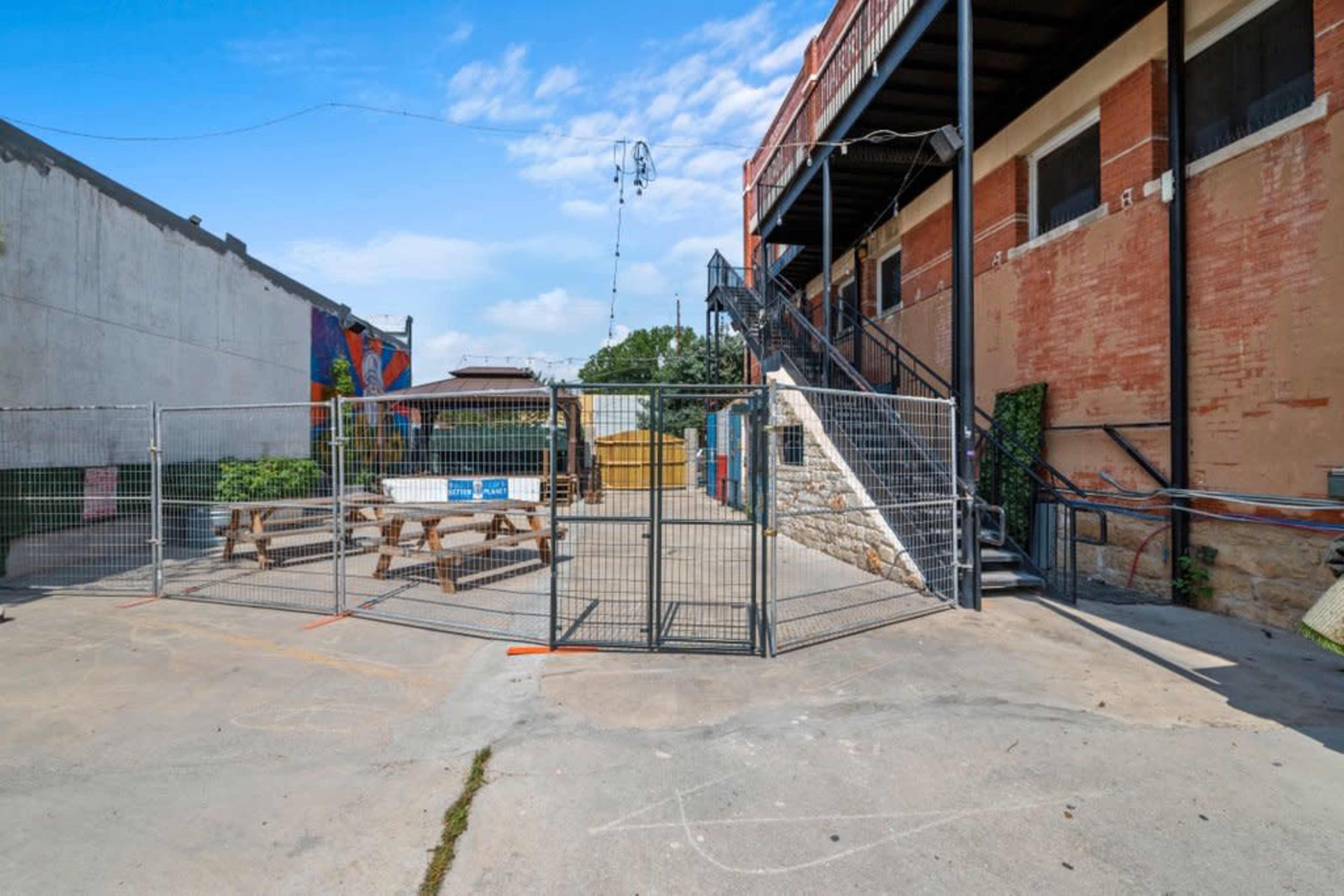 The image shows a fenced area with picnic tables, located beside a brick building, with a staircase leading to an upper level.