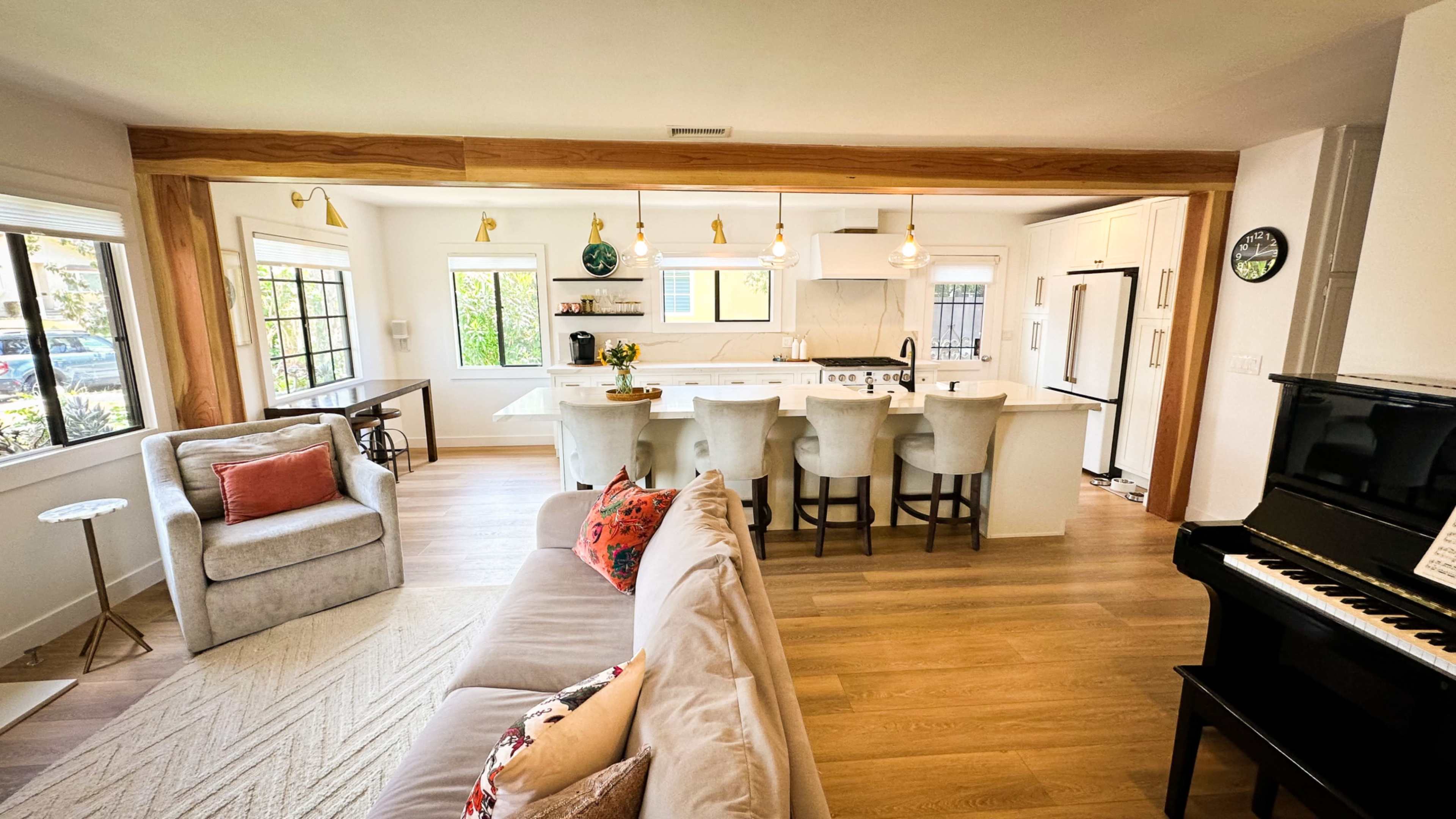 The image shows an open-concept kitchen and living area, featuring a modern kitchen with a large island, bar stools, and a piano in the corner of the living space.