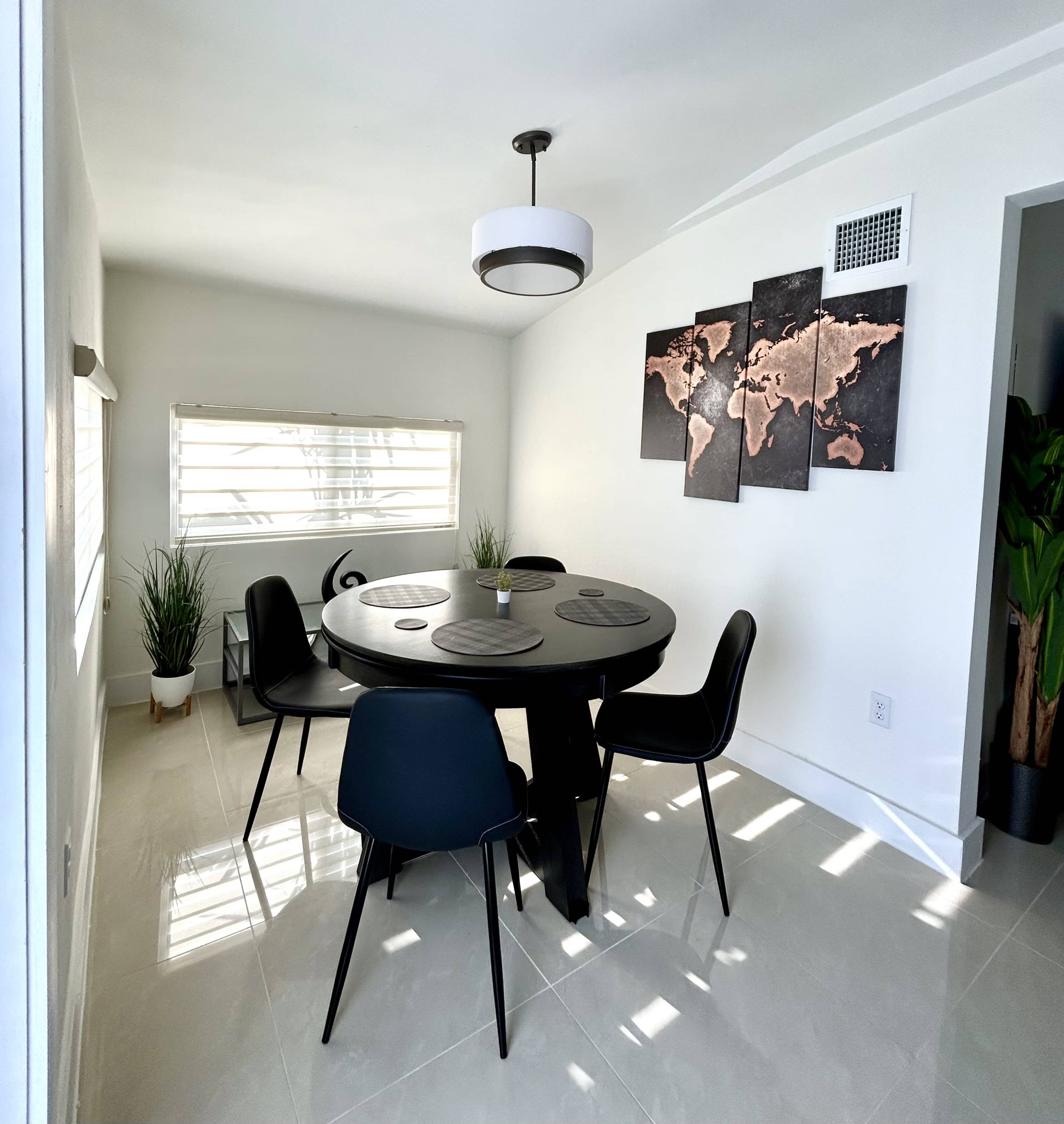 A modern dining room features a round black table surrounded by four black chairs, with a world map art piece on the wall and plants in the corners.