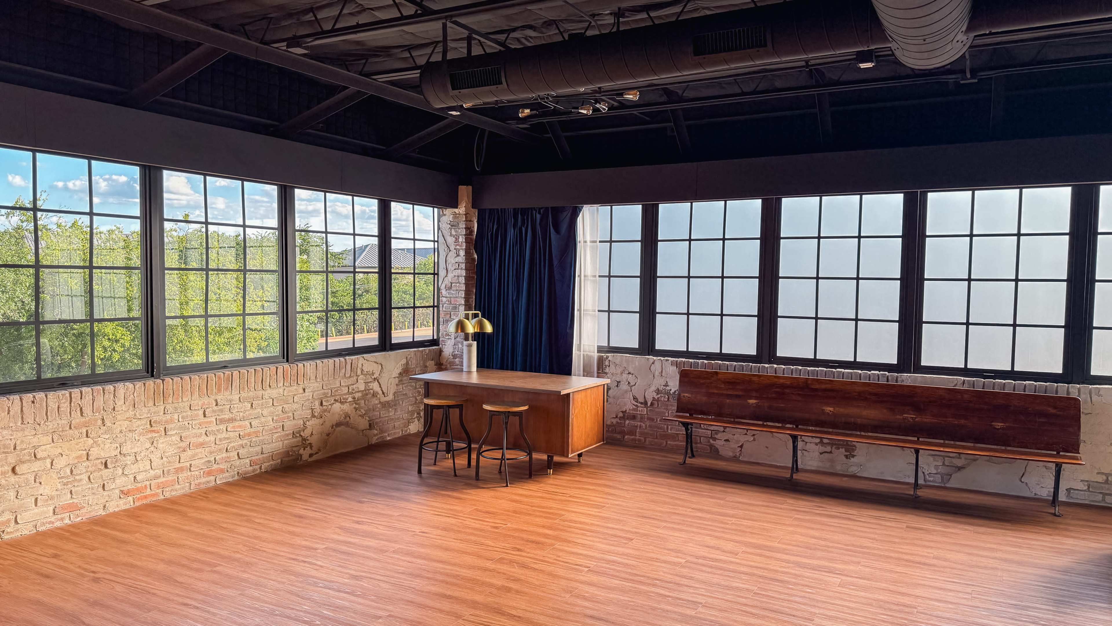 The image shows a spacious room with large windows, a wooden table with two stools, and a wooden bench against the wall.