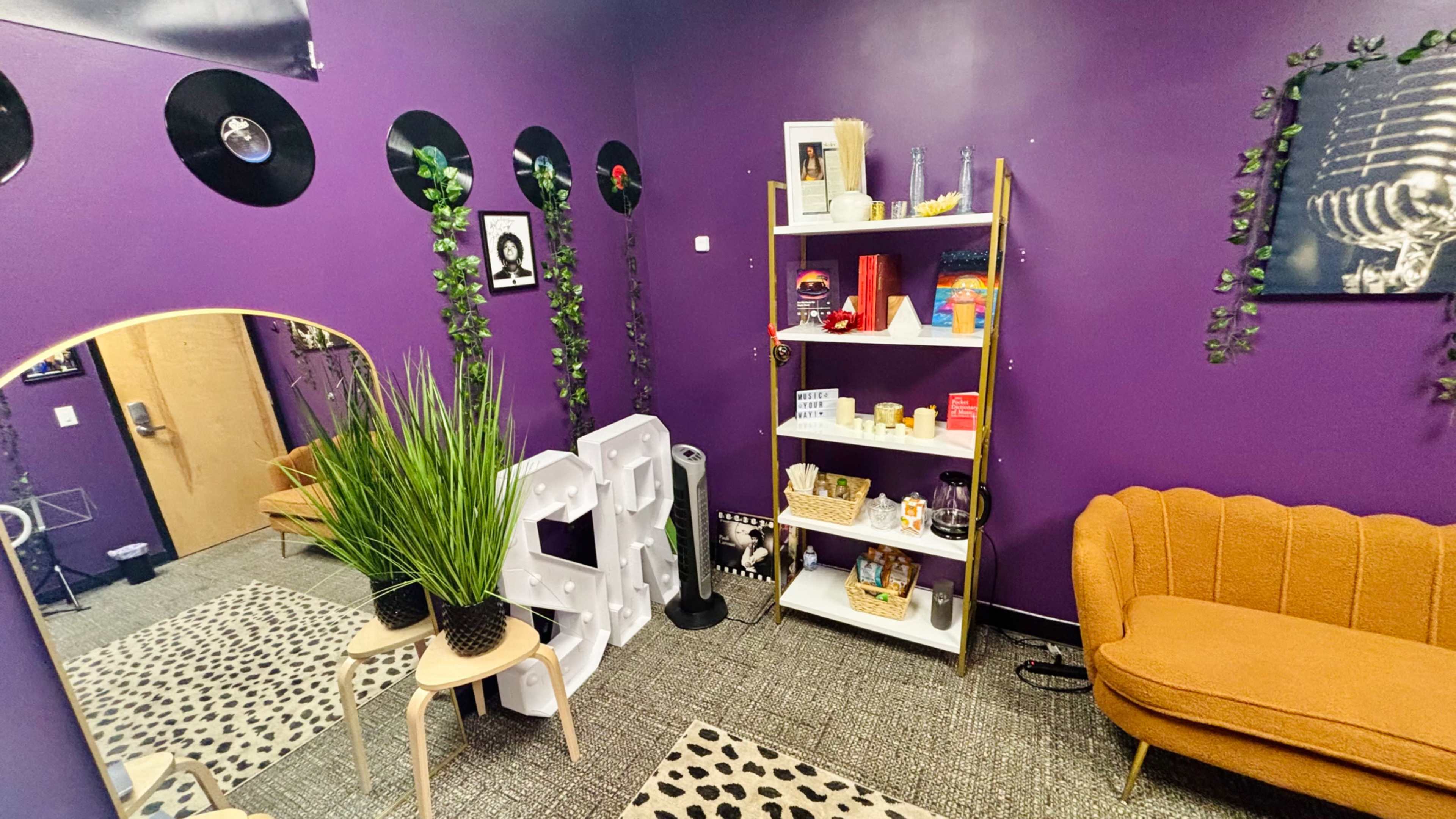The room features purple walls adorned with vinyl records, a cozy orange couch, a shelving unit filled with various decorative items, and a large plant, all set on a patterned carpet.
