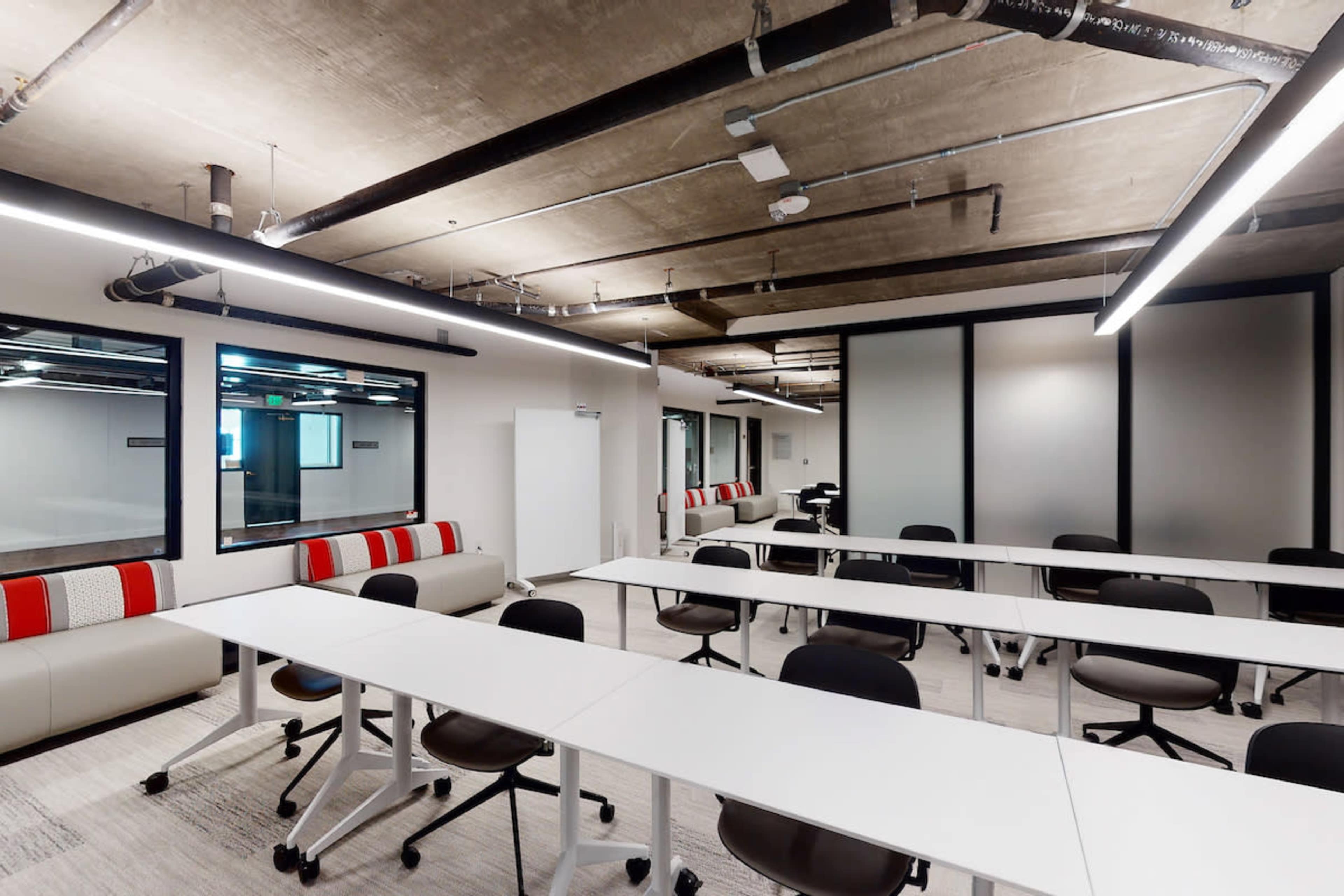 A modern office space with rows of white tables and chairs, and large windows along the walls.