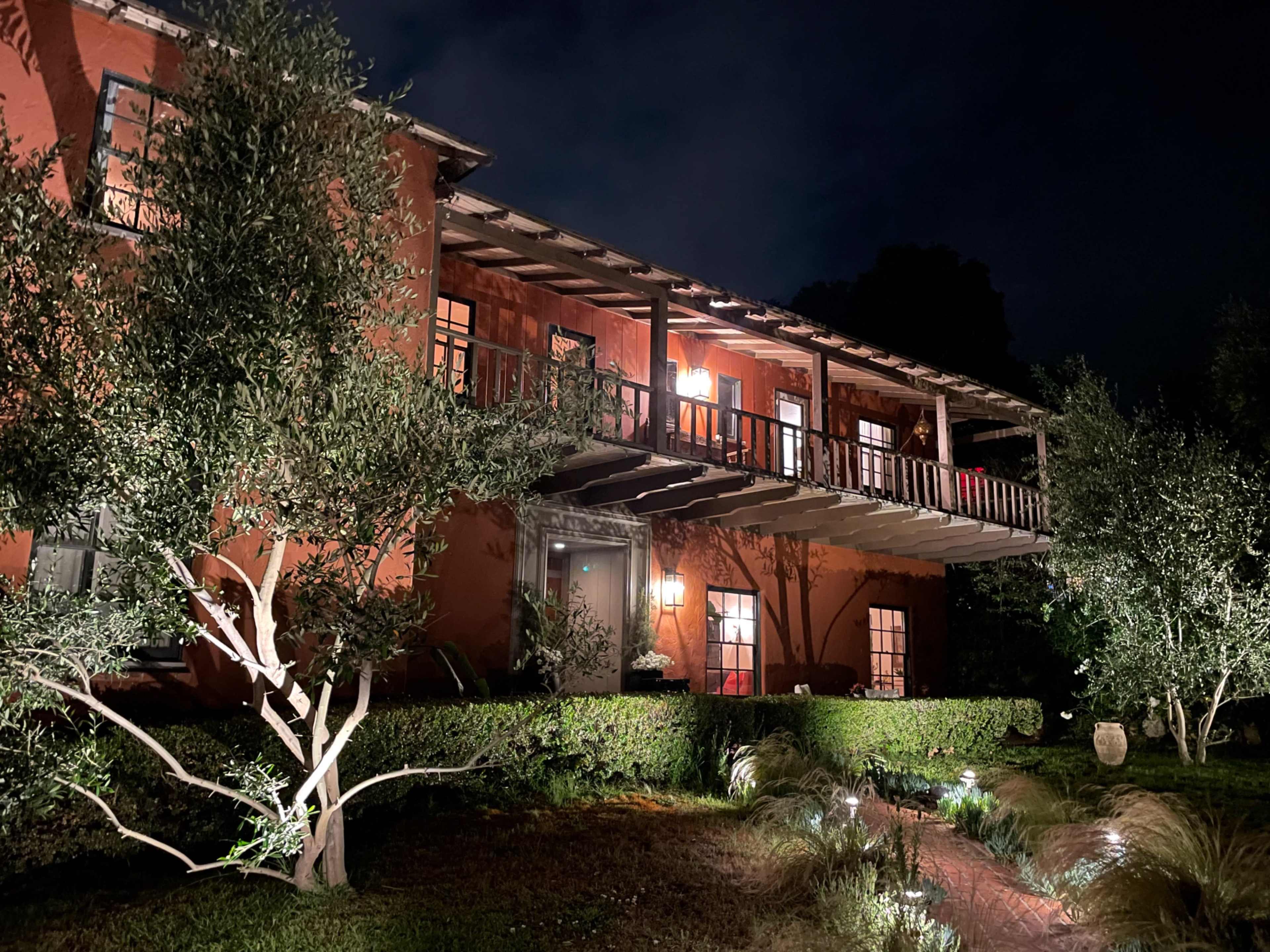 A two-story building with a balcony, surrounded by trees and illuminated by outdoor lights, is set against a dark night sky.