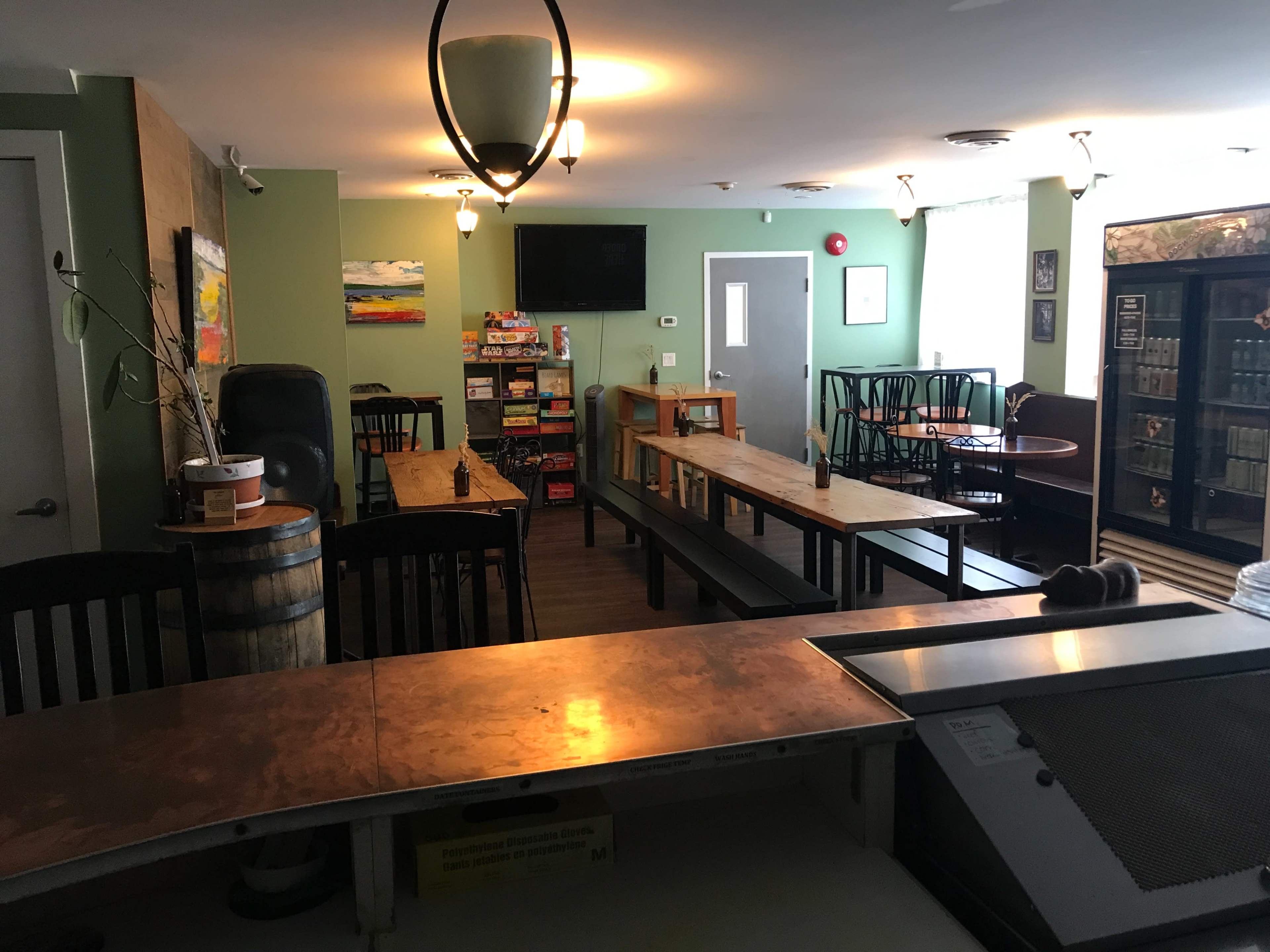 A spacious, empty café with wooden tables, chairs, and a counter, featuring shelves of beverages and games in the background.